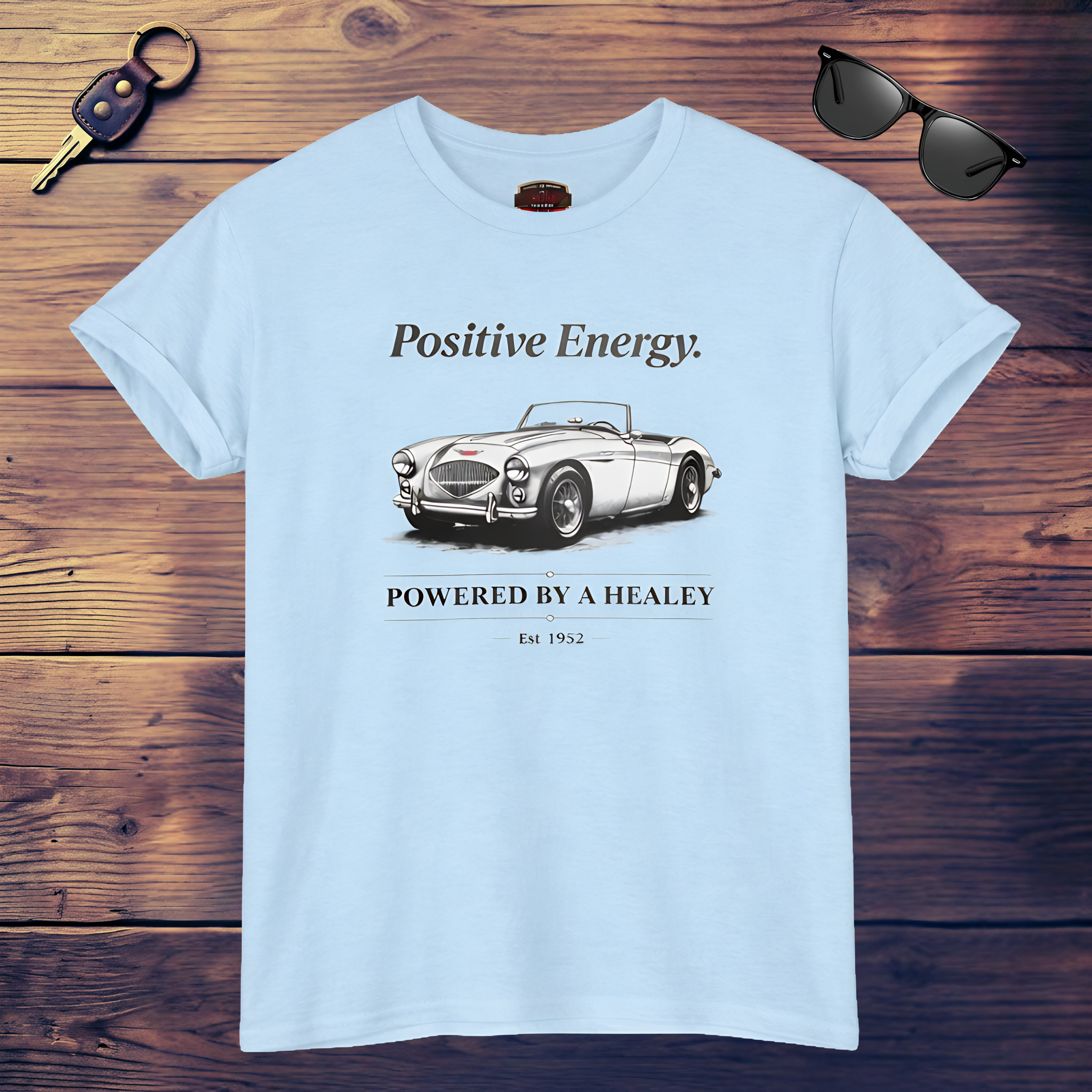 White t-shirt with a vintage car graphic and text on a wooden surface, accompanied by sunglasses and a key.