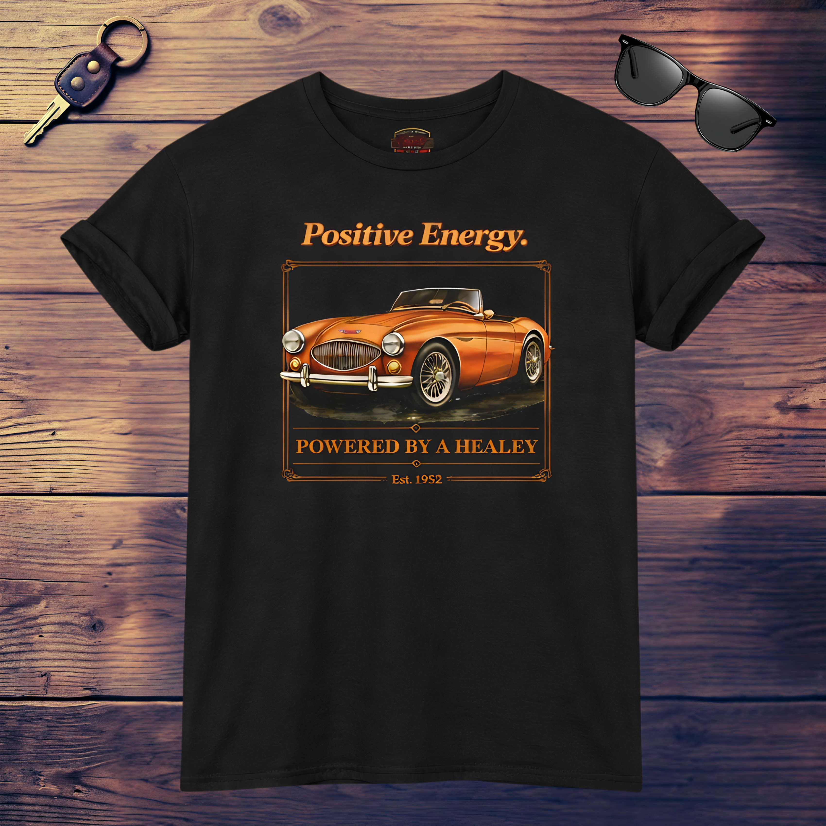 Black t-shirt with orange classic car graphic and text on a wooden surface with sunglasses and keys.