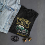 Black t-shirt with 'Never Underestimate a Dad with a Healey' design, denim jacket, sunglasses, and beads on a gray surface.