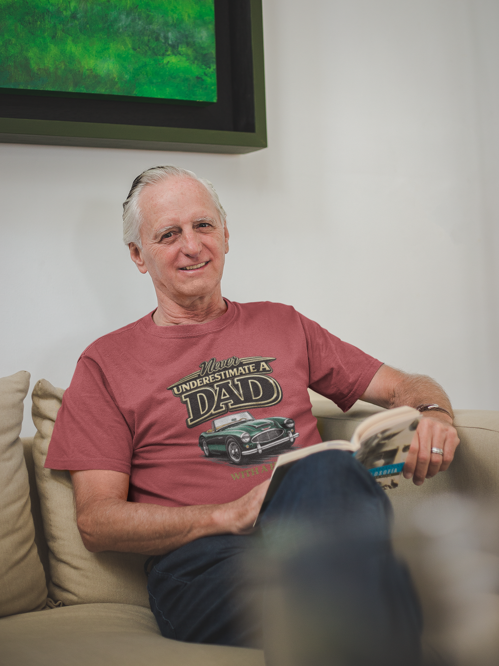 Man sitting on a couch wearing a heather red t-shirt with text and classic car graphics.