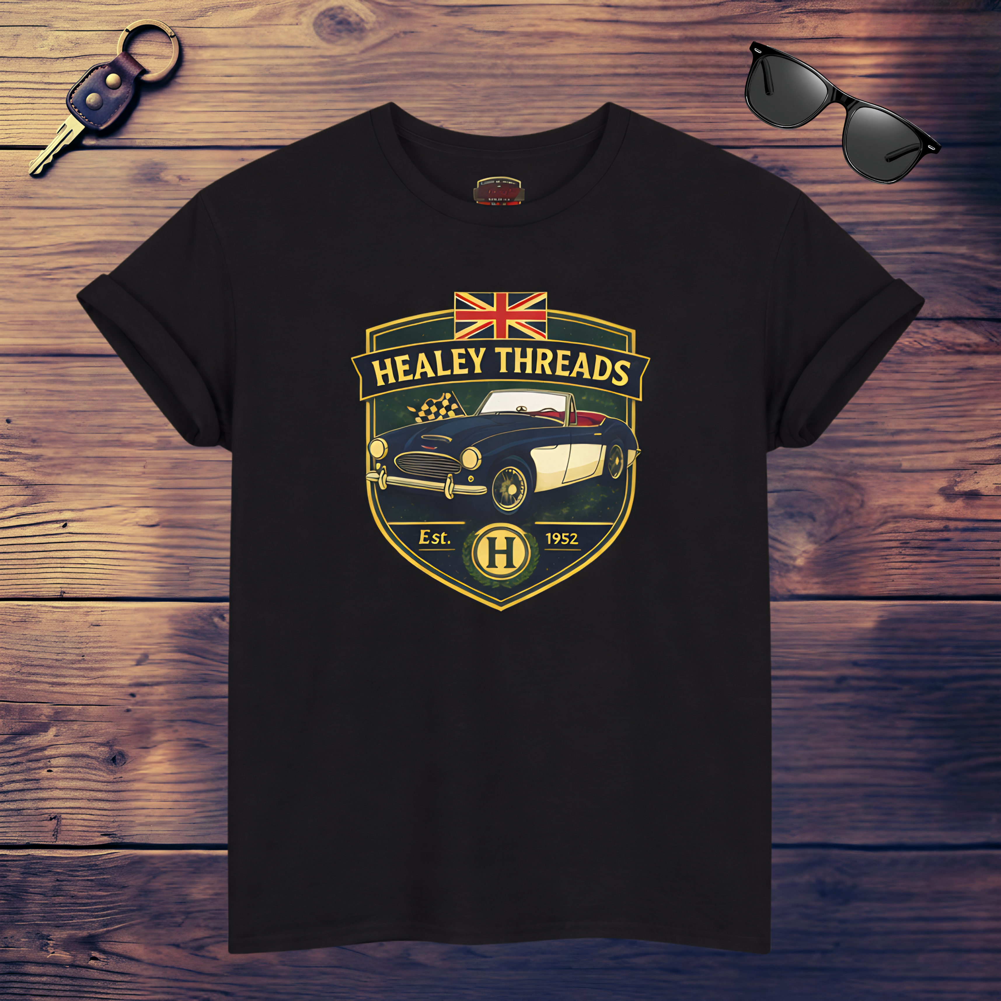 Black t-shirt with 'Healey Threads' logo on a wooden surface with sunglasses and keys.