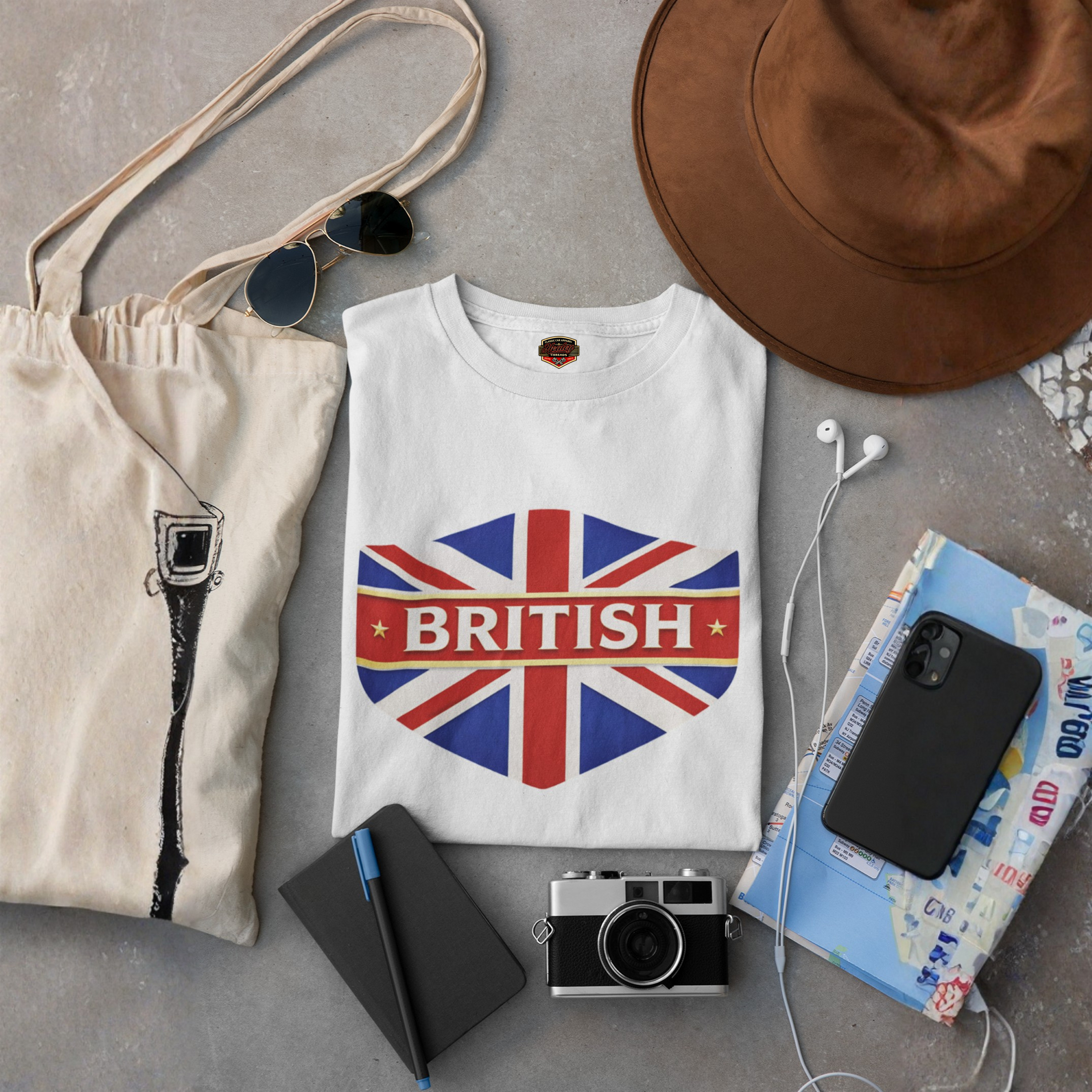 White t-shirt with British flag design, beige tote bag, sunglasses, camera, map, and phone on a gray surface.