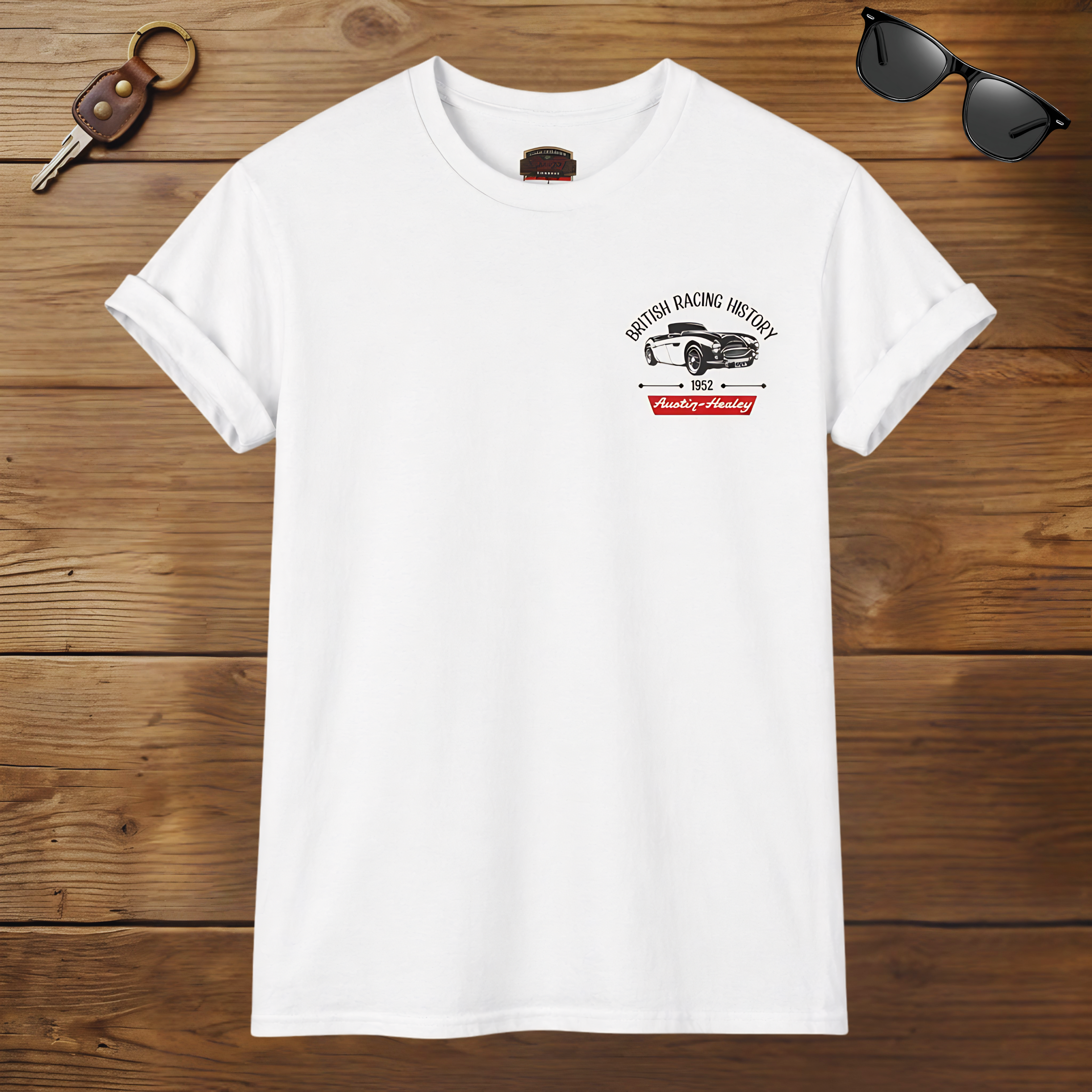 White t-shirt with a graphic Healey design on a wooden surface with sunglasses and keys.