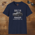 Navy blue t-shirt with car graphic and text on a wooden surface with sunglasses and keys.