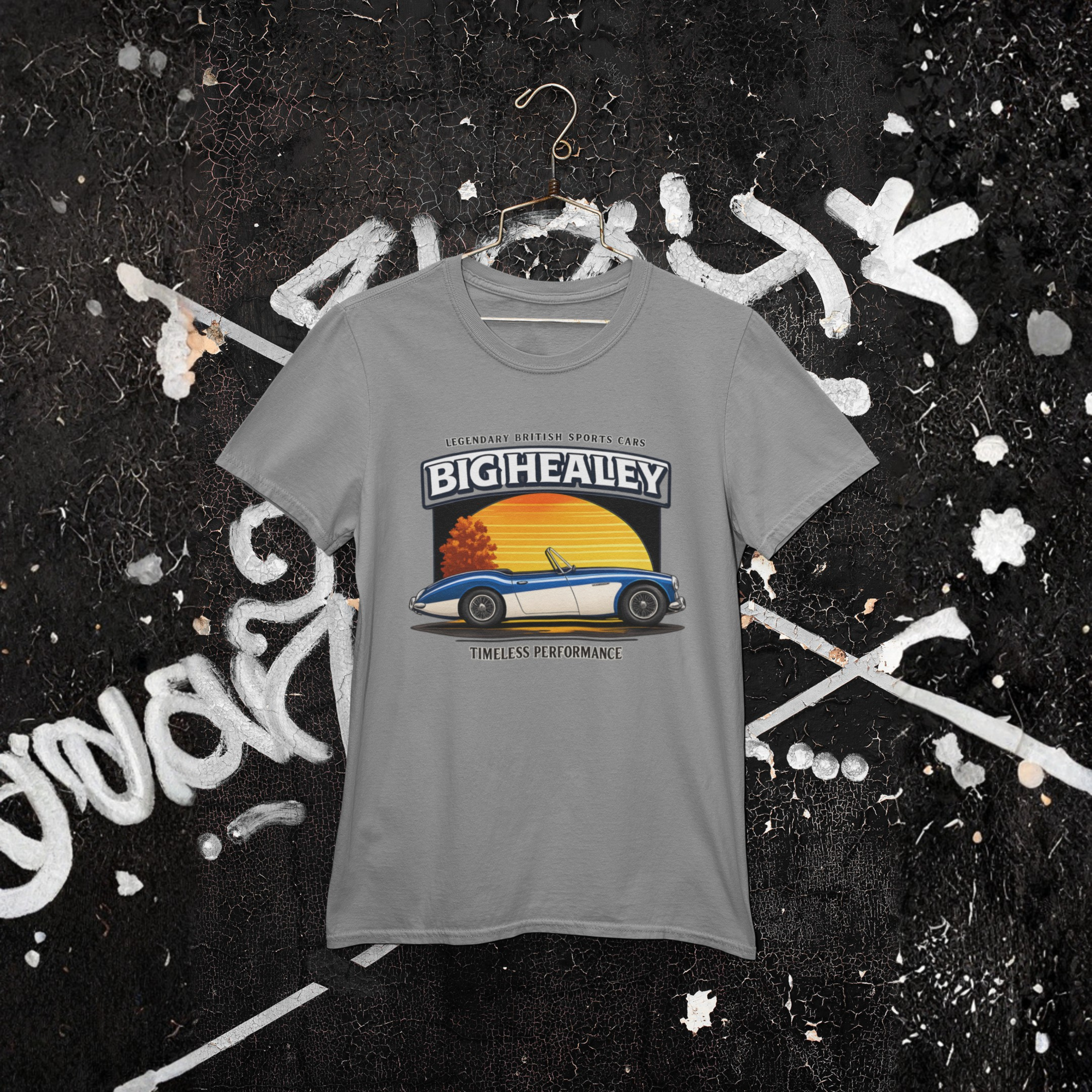 Big Healey graphic gray colored t-shirt featuring a blue and white Austin Healey roadster with retro sunset artwork and vintage British sports car hanging against a wall.