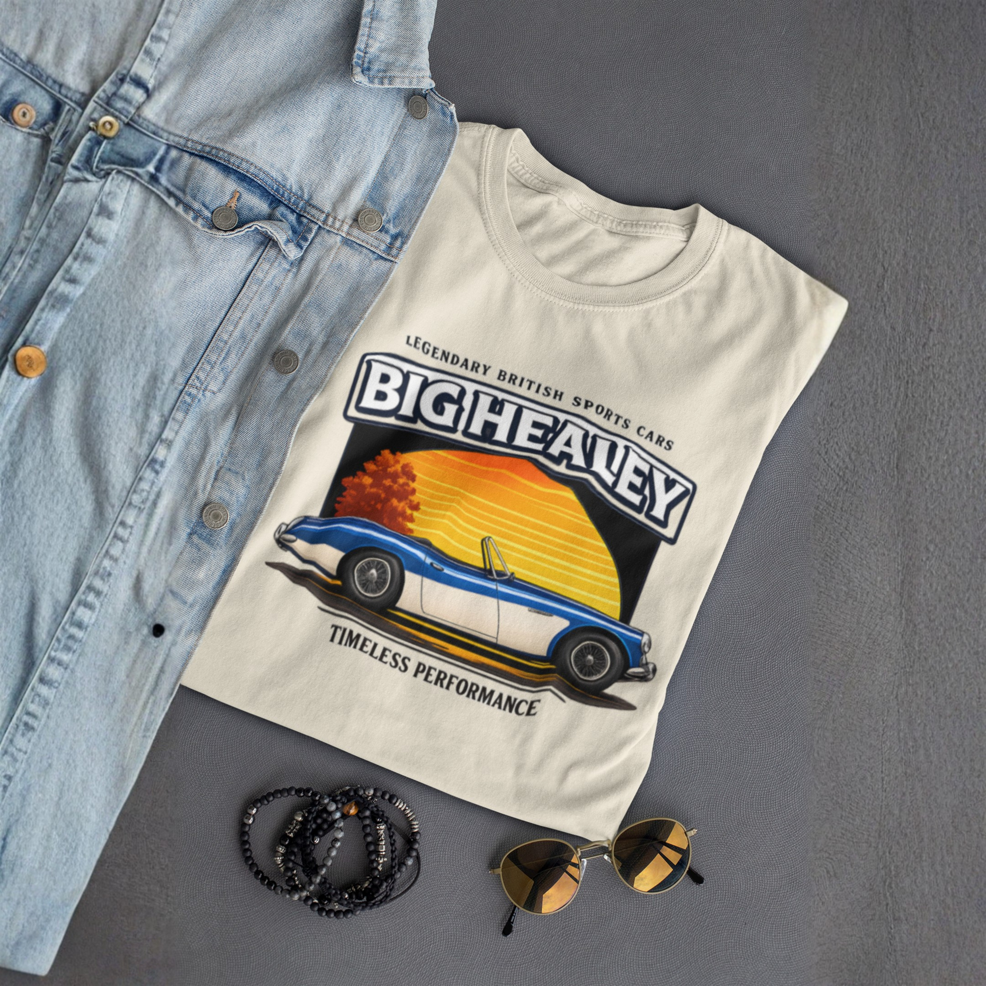 Folded Big Healey graphic natural colored t-shirt featuring a blue and white Austin Healey roadster with retro sunset artwork and vintage British sports car on a gray background