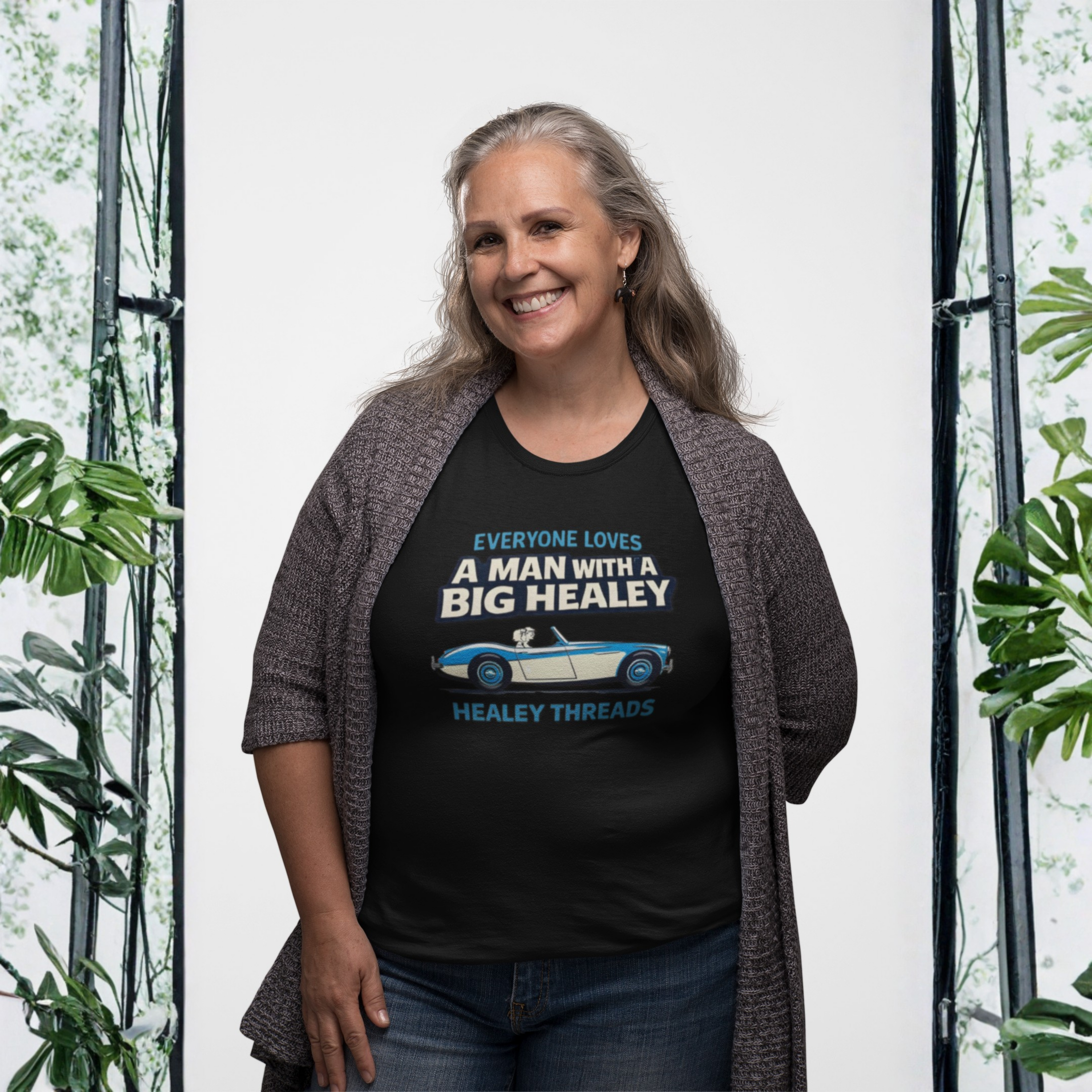 Woman wearing a black t-shirt with a Healey graphic and text, standing in front of a white background with plants.