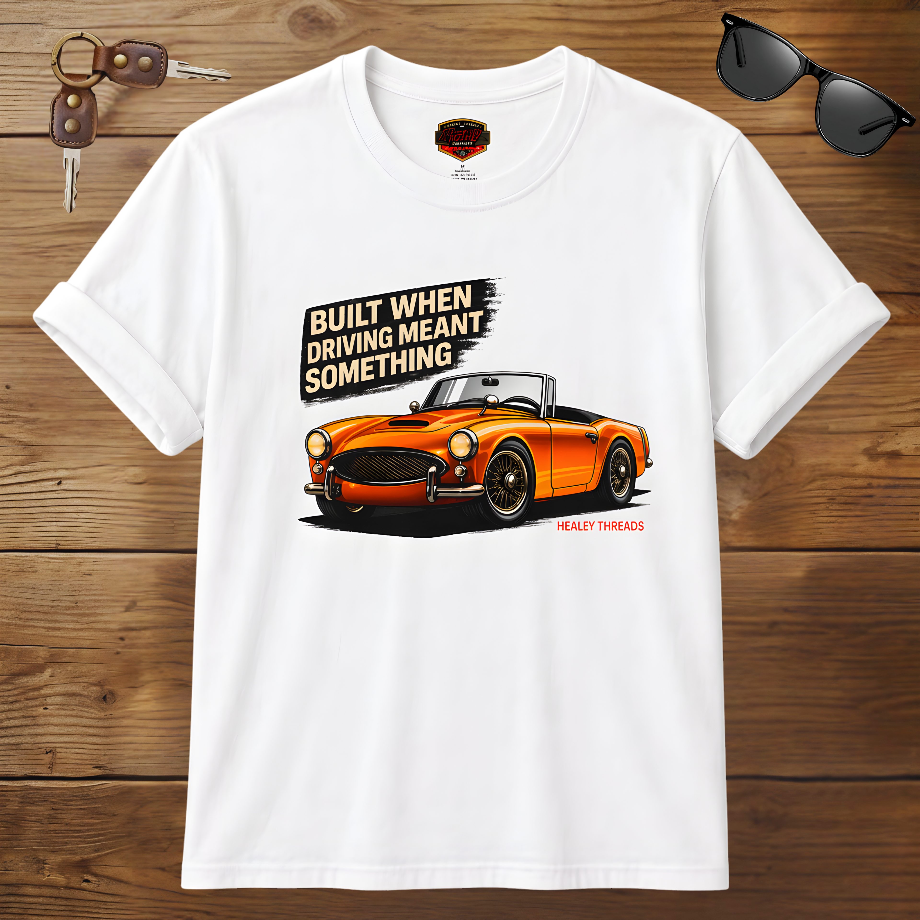 White t-shirt with orange car graphic and text on a wooden surface with sunglasses and keys.