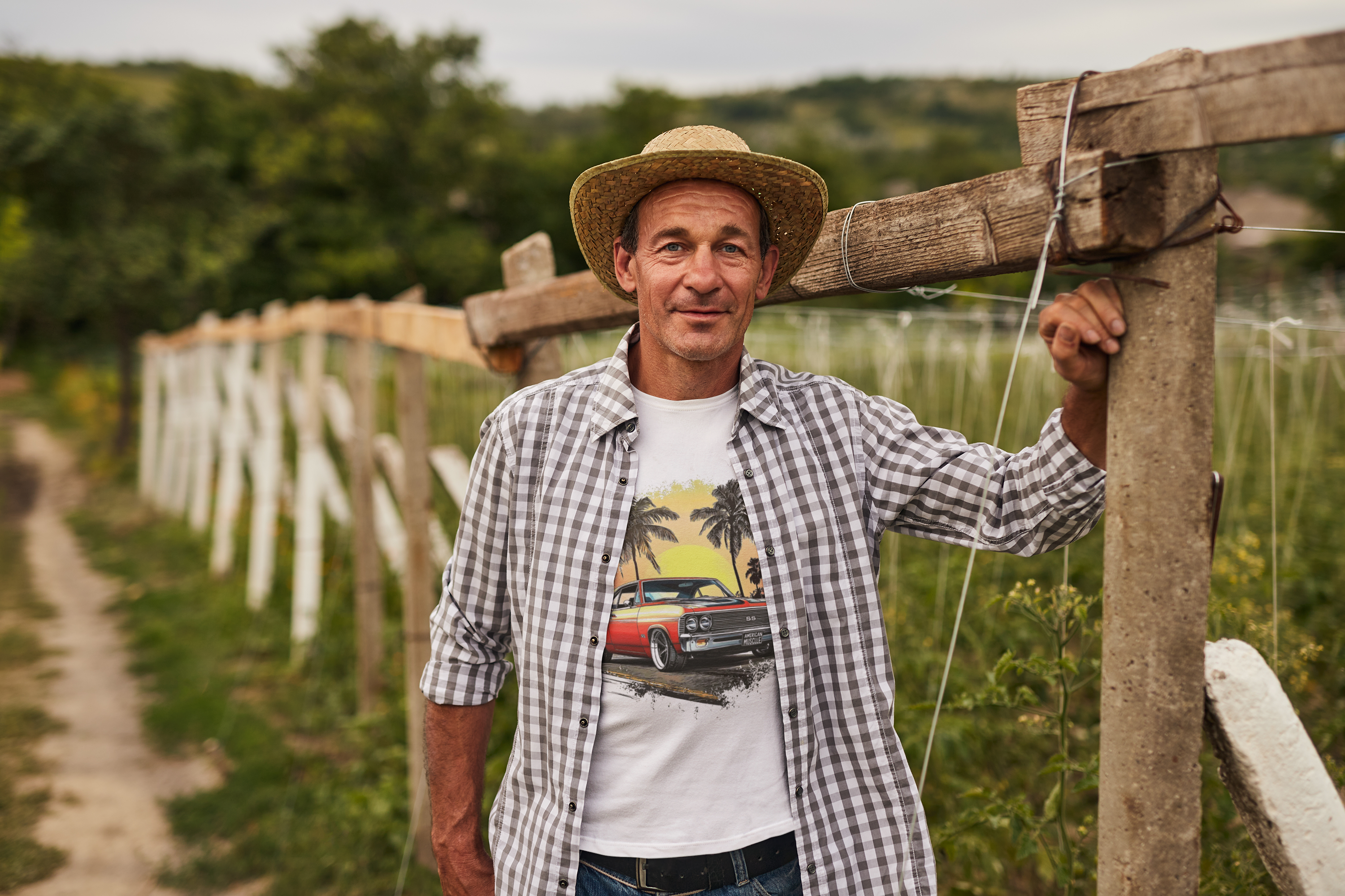 Man wearing a checkered shirt with a graphic classic car design, standing next to a wooden fence in a rural setting.