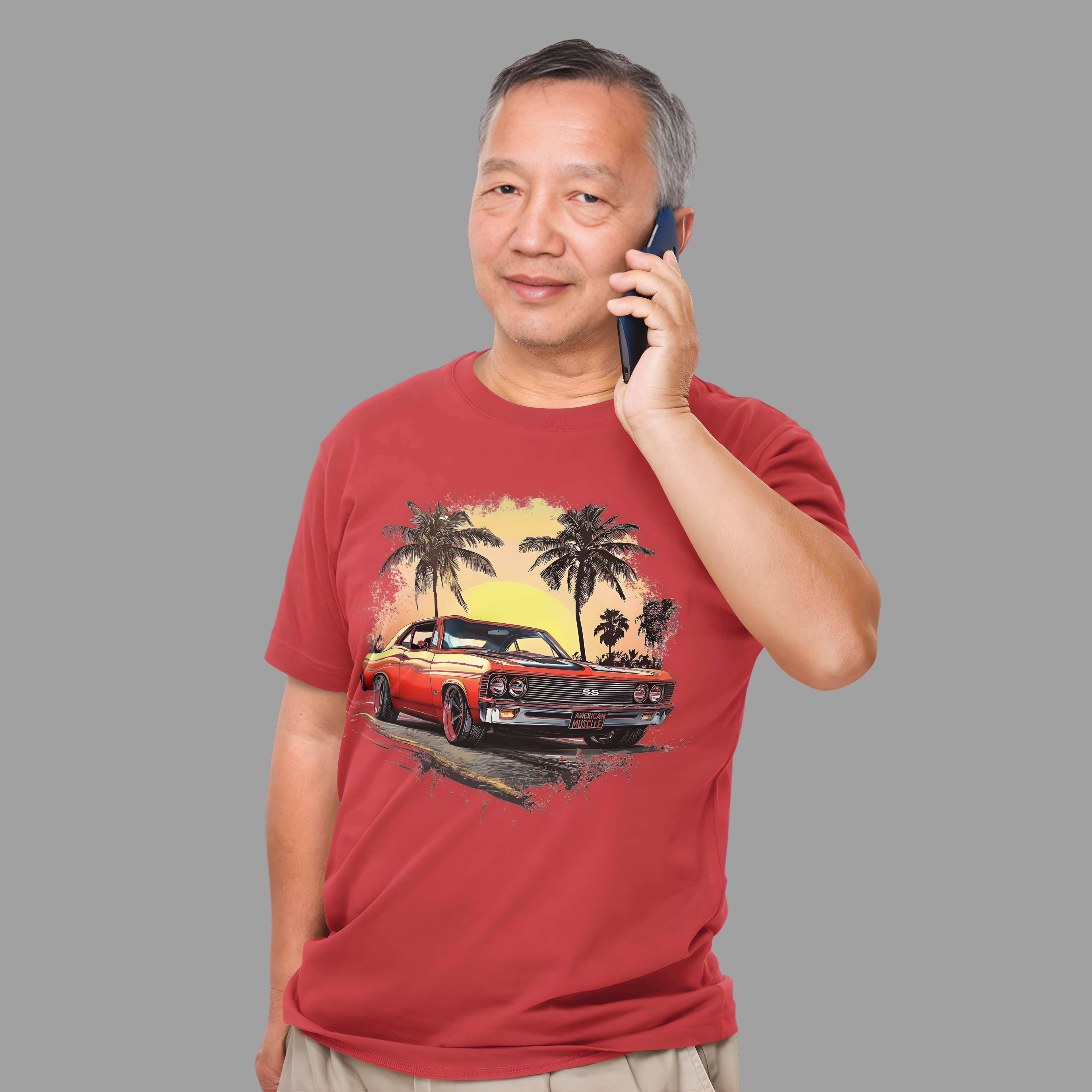 Man wearing a red t-shirt with a classic car and palm tree design, talking on a phone against a gray background