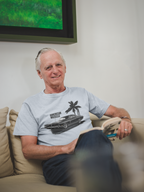Man sitting on a couch wearing a gray t-shirt with a graphic design, with a television in the background.