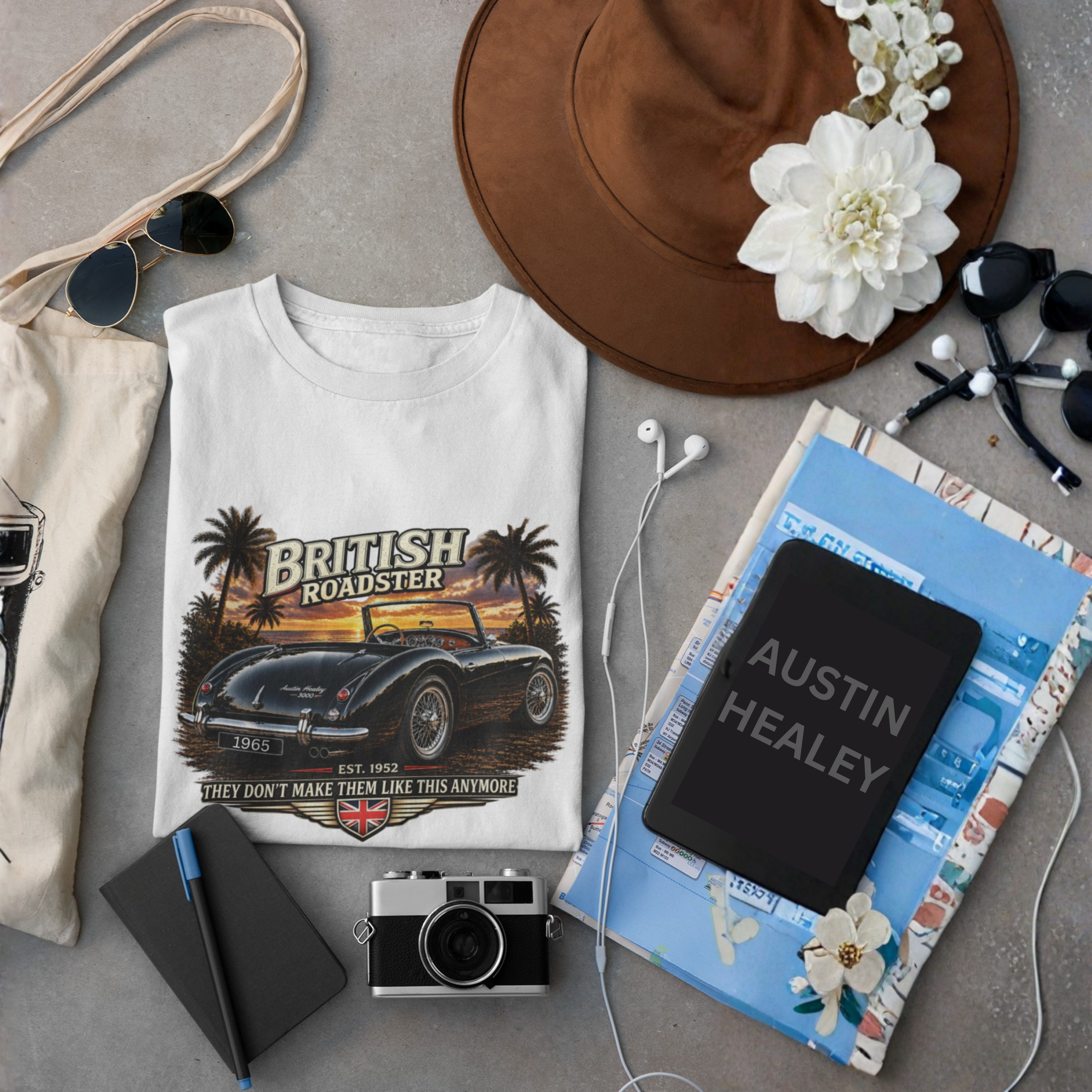 Collection of items including a white t-shirt with a British Roadster design, notebook, camera, and accessories on a gray surface.
