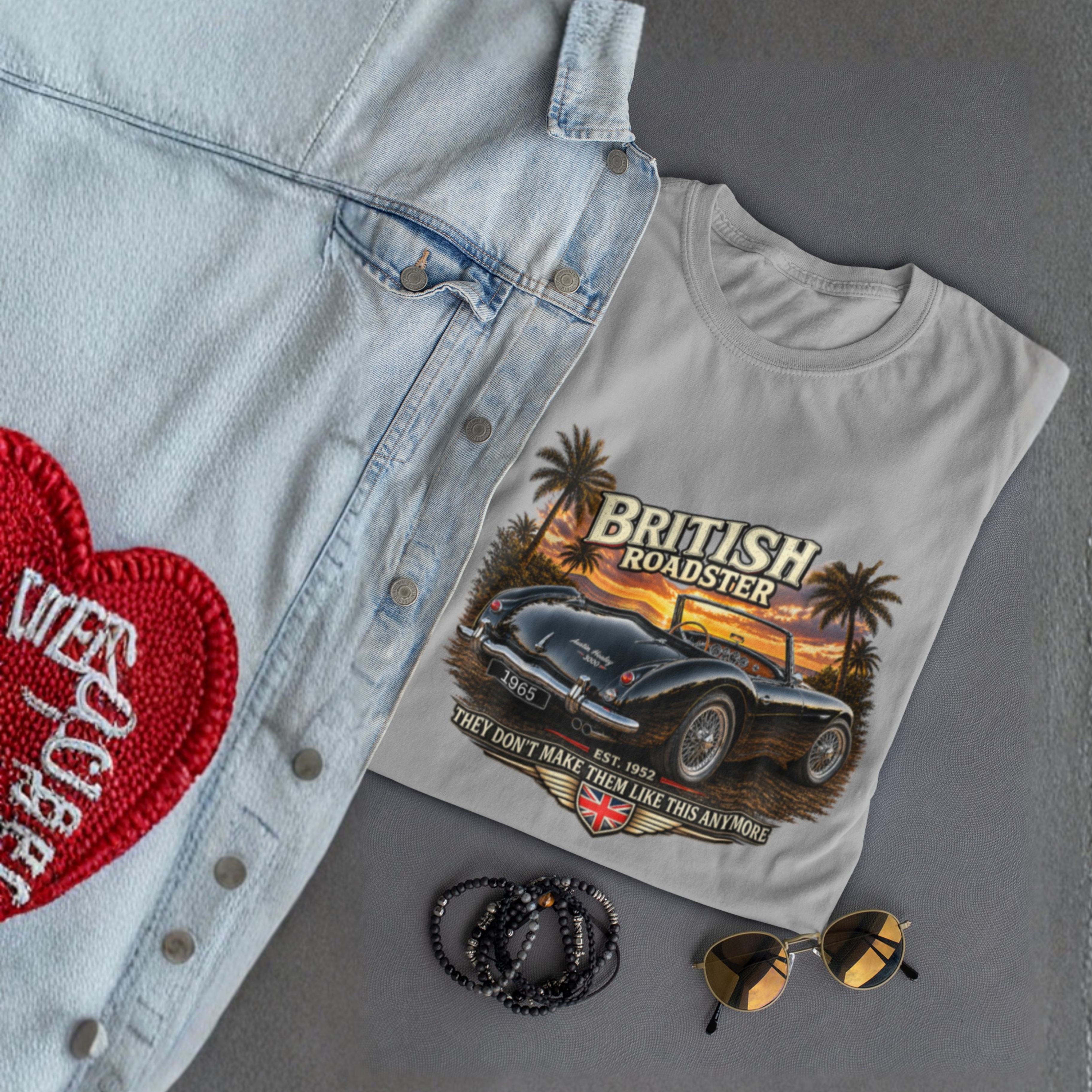 Sport gray t-shirt with 'British Roadster' graphic design, denim jacket with red heart patch, sunglasses, and necklace on gray surface.