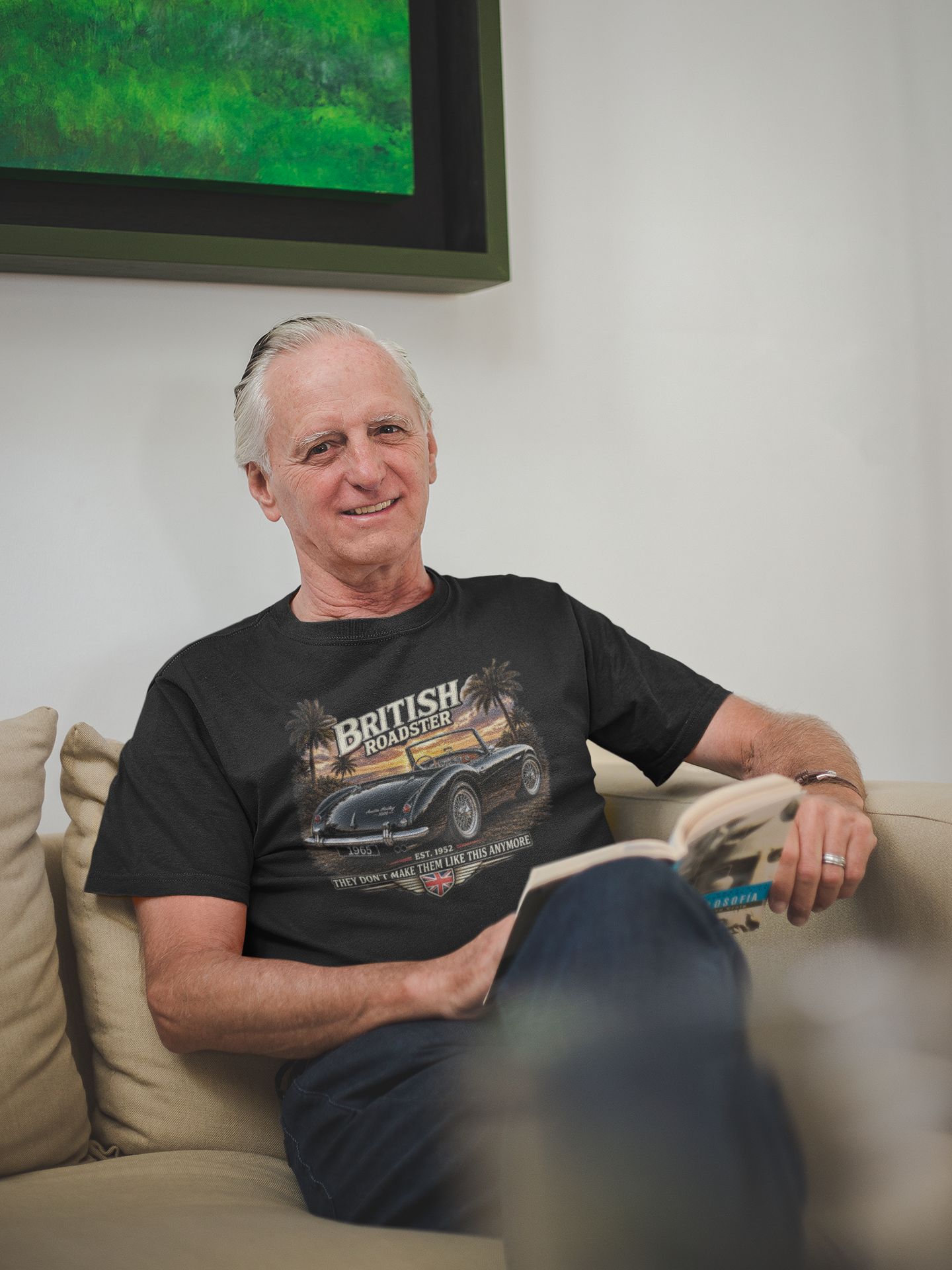 Man sitting on a couch wearing a black t-shirt with a graphic design, in a room with a painting on the wall.