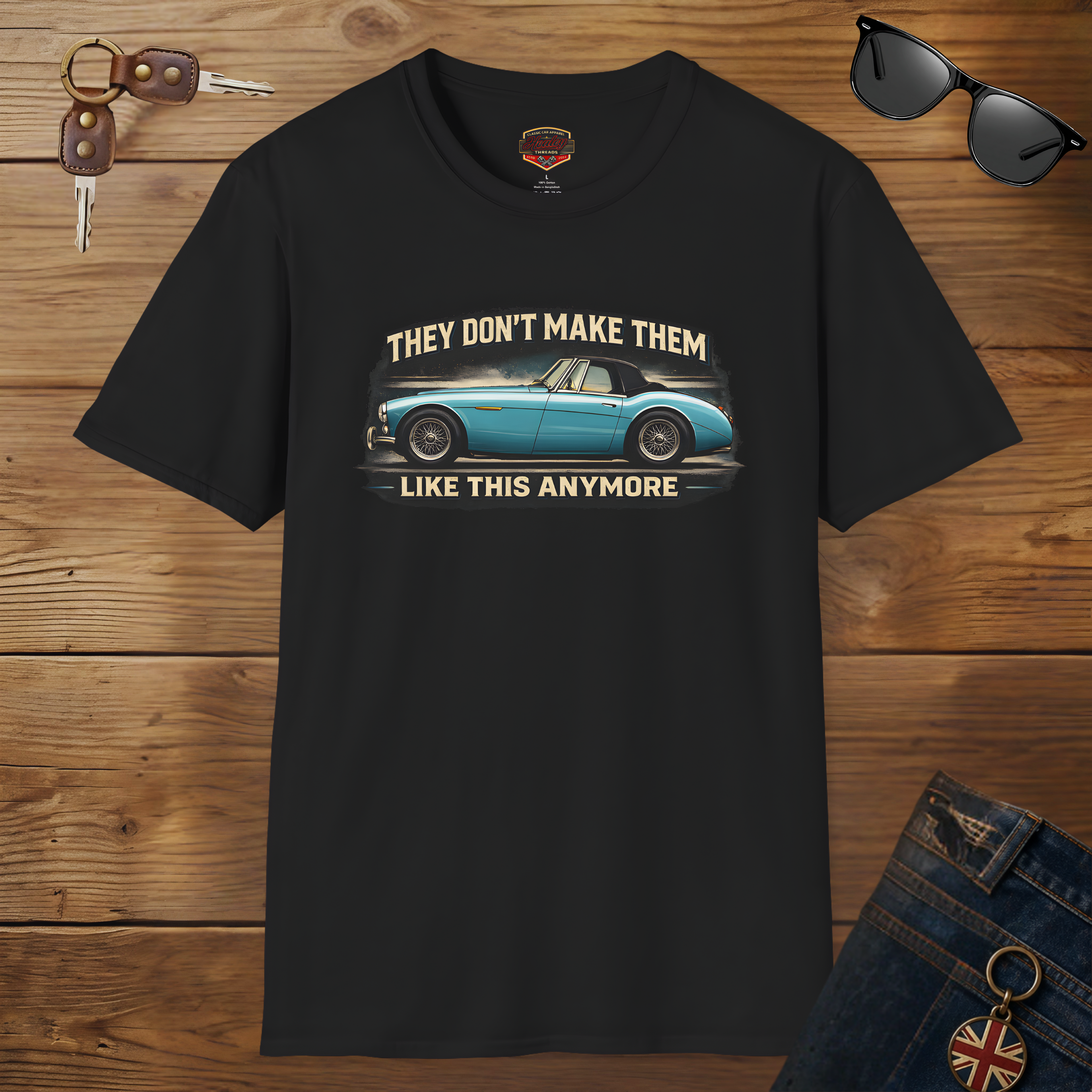 Black t-shirt with a graphic of a classic car and text on a wooden surface with sunglasses and keys.