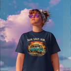 Woman with wavy hair wearing a denim jacket over a white t-shirt against a blue sky, featuring a retro surf van graphic reading "Sun. Salt. Air., Coastal Cruisers, Healey Threads."