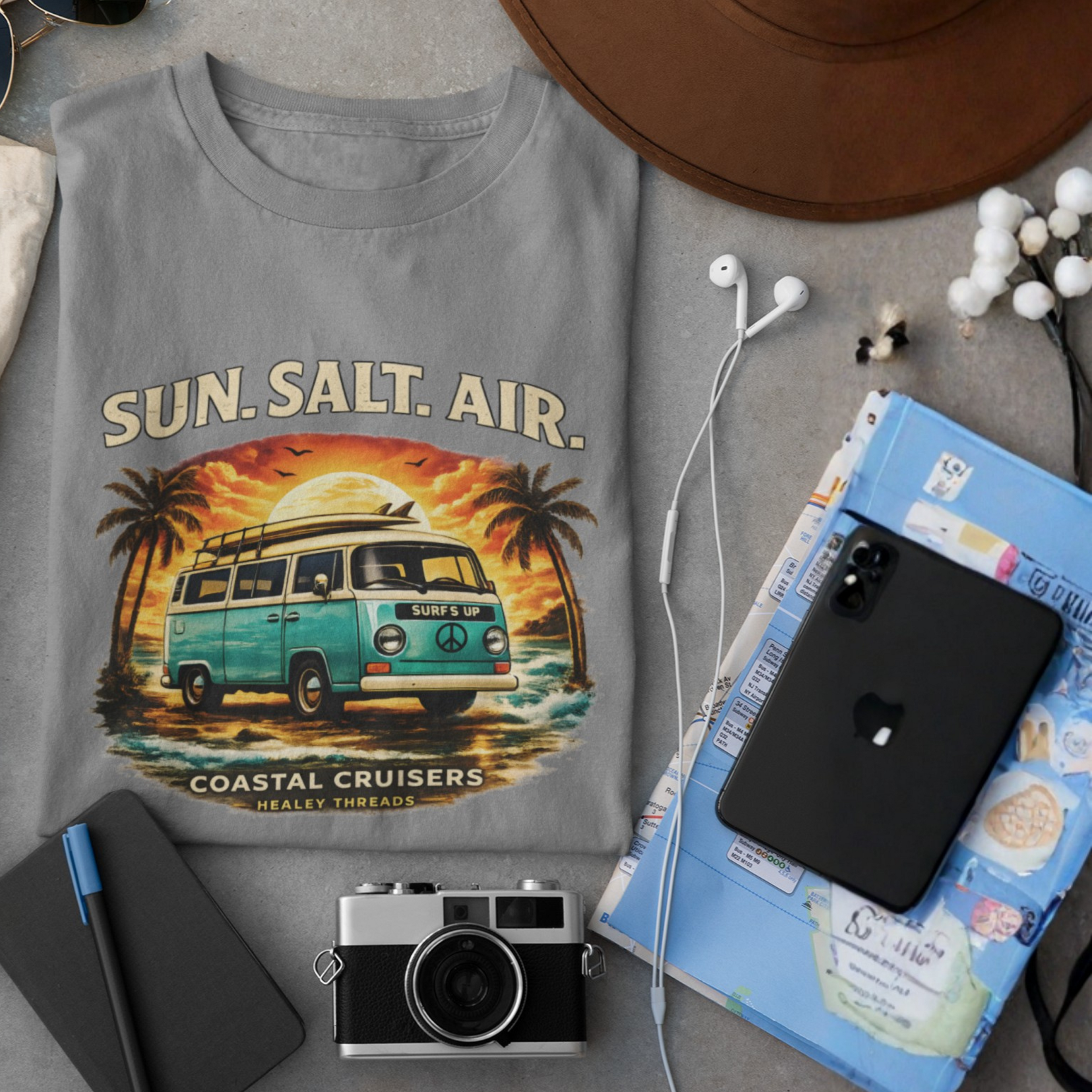 Gray t-shirt with a van and palm trees design labeled 'Sun. Salt. Air.' on a table with a phone, camera, and notebook.