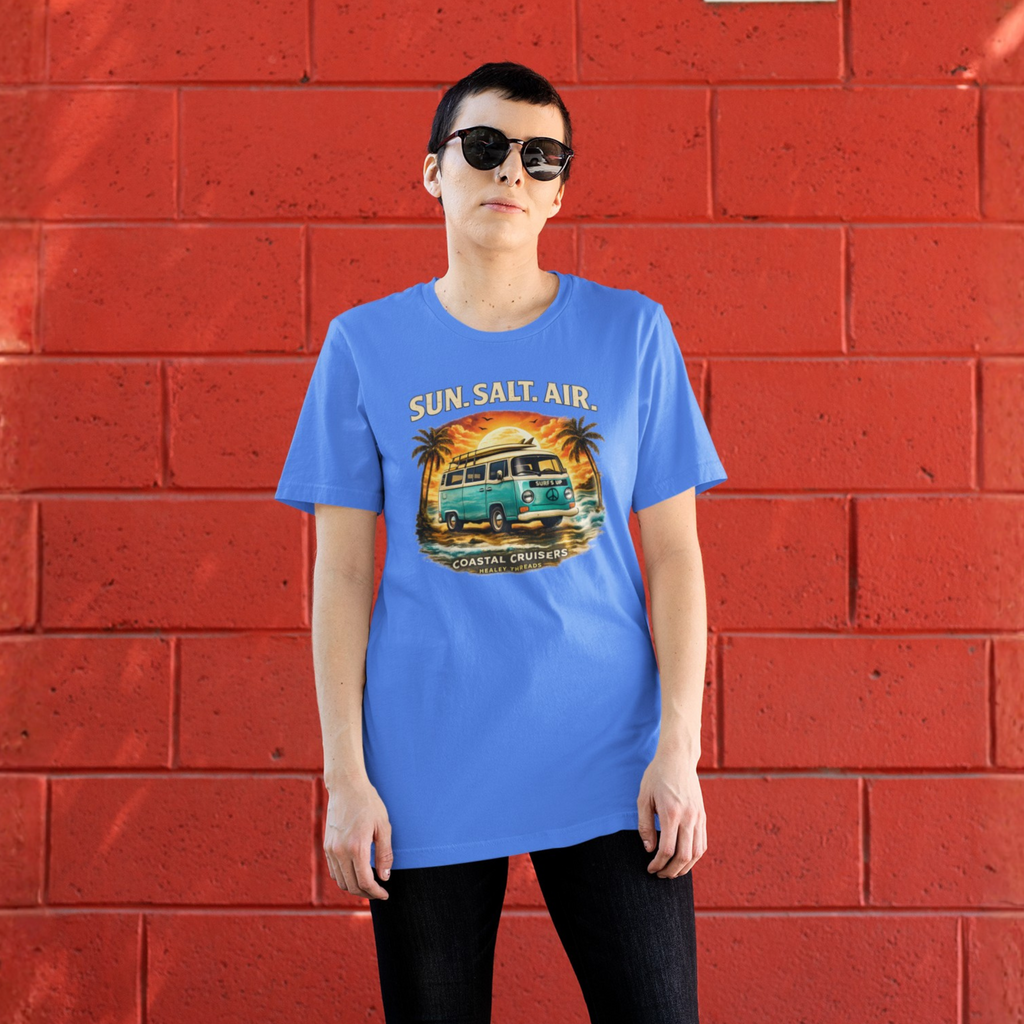 Woman wearing a blue retro coastal cruiser T-shirt featuring a vintage turquoise surf van, palm trees, ocean waves, and a sunset, standing in front of a bright red brick wall.