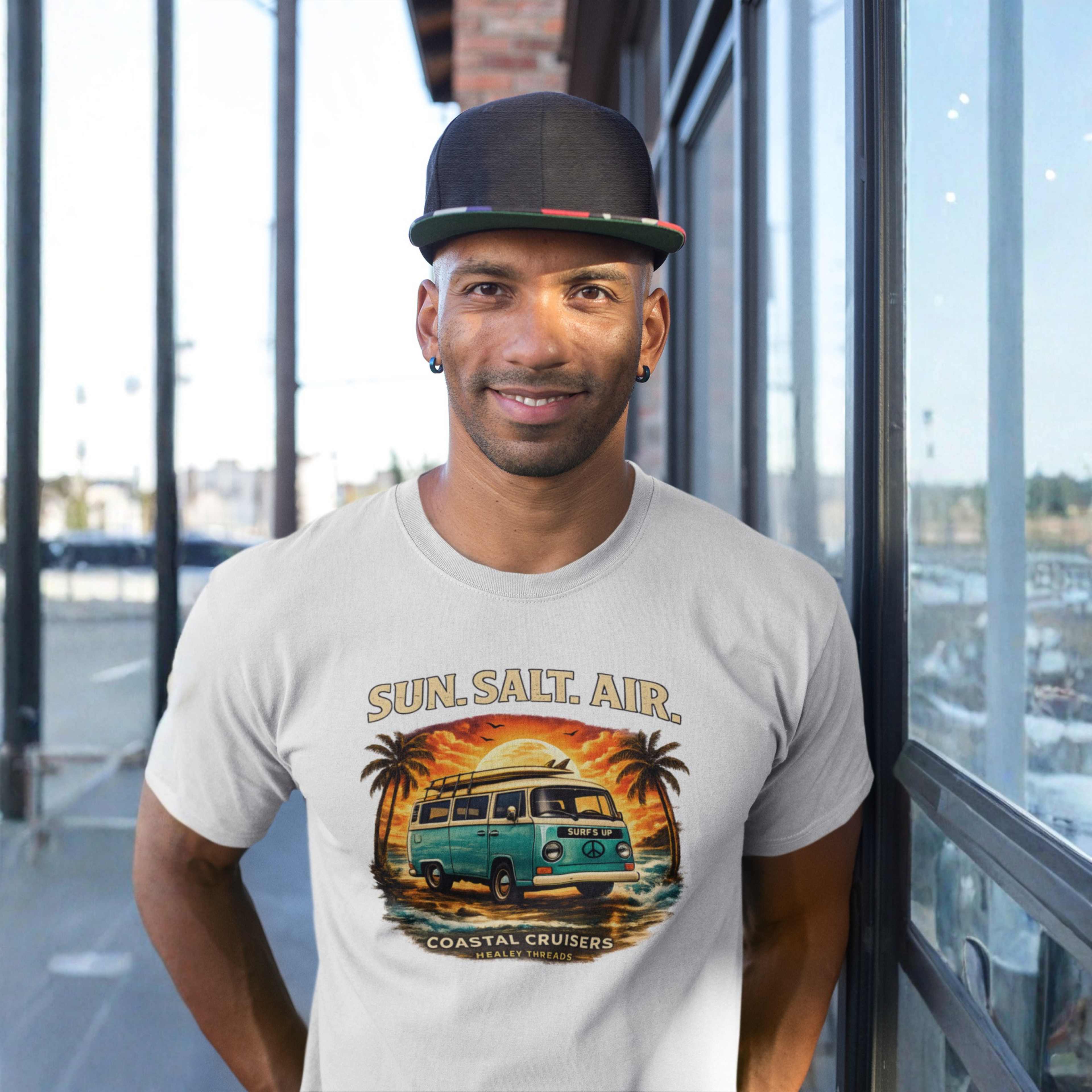 Smiling man wearing a black cap and light gray t-shirt standing on an outdoor walkway, featuring a retro surf van graphic reading "Sun. Salt. Air., Coastal Cruisers, Healey Threads."