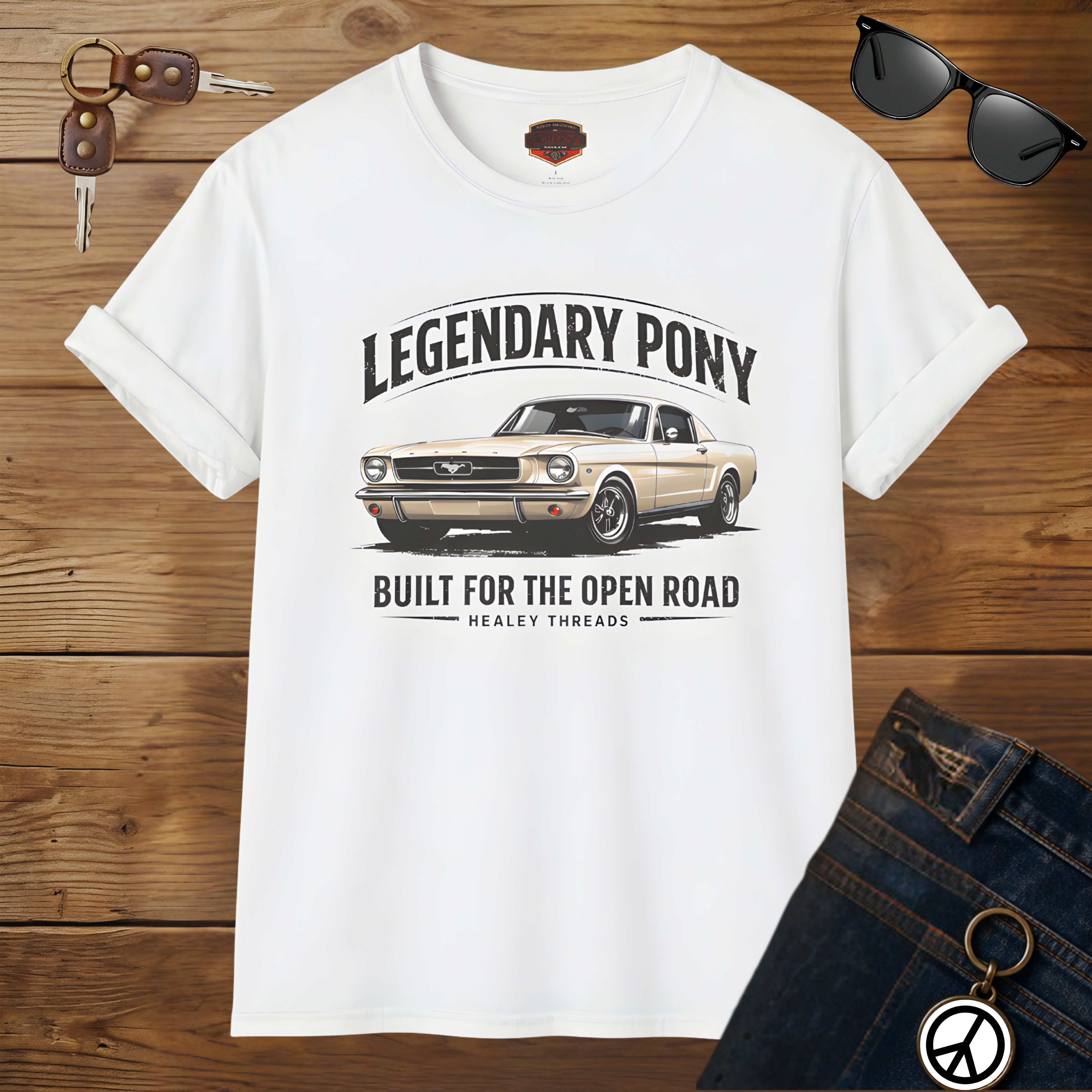 White t-shirt with car graphic and text on a wooden surface with sunglasses and keys.