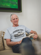 Man sitting on a couch wearing a sport gray t-shirt with a graphic classic car design, with a television in the background. 