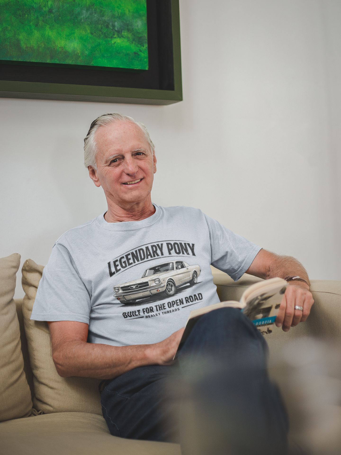Man sitting on a couch wearing a sport gray t-shirt with a graphic classic car design, with a television in the background. 