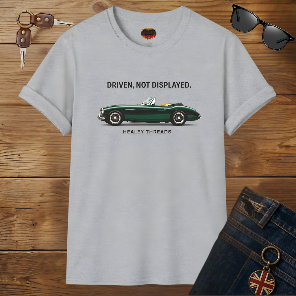 Sport gray t-shirt with a green classic car graphic and text on a wooden surface, surrounded by sunglasses, keys, and jeans.