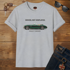 Sport gray t-shirt with a green classic car graphic and text on a wooden surface, surrounded by sunglasses, keys, and jeans.