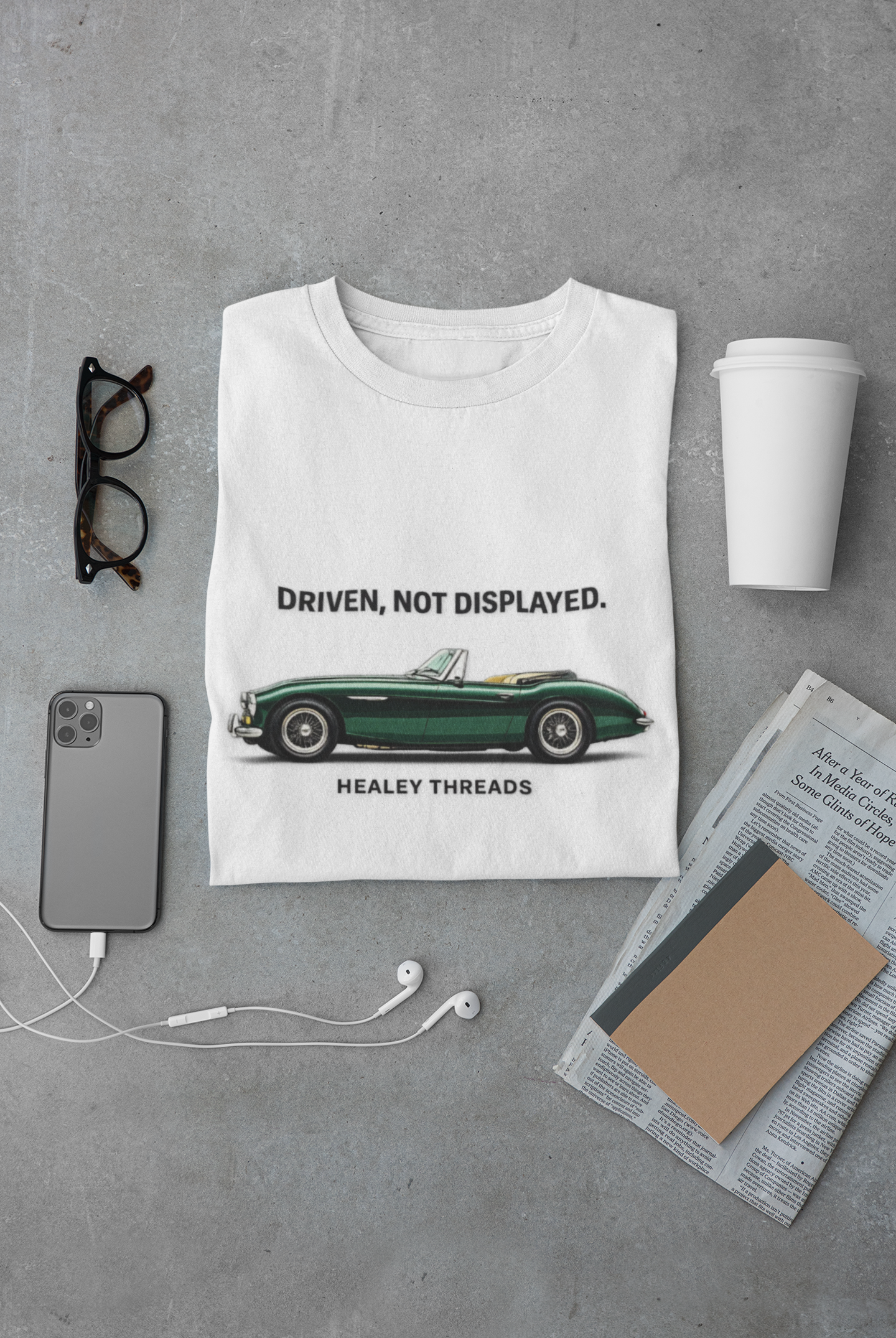Folded white t-shirt with vintage car graphic and text on a gray surface with accessories.