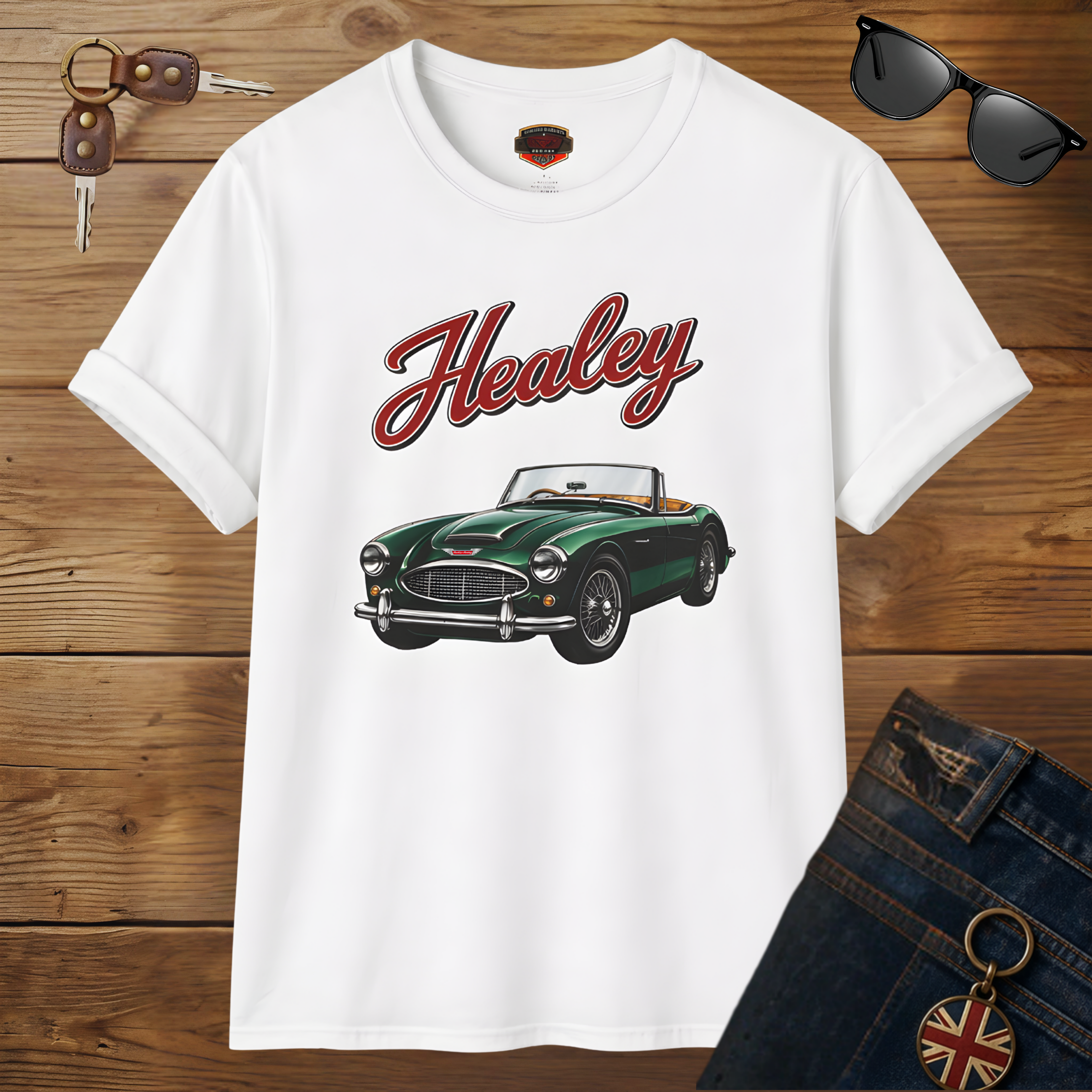 White t-shirt with green classic car graphic and 'Healey' text on a wooden surface with sunglasses and keys.