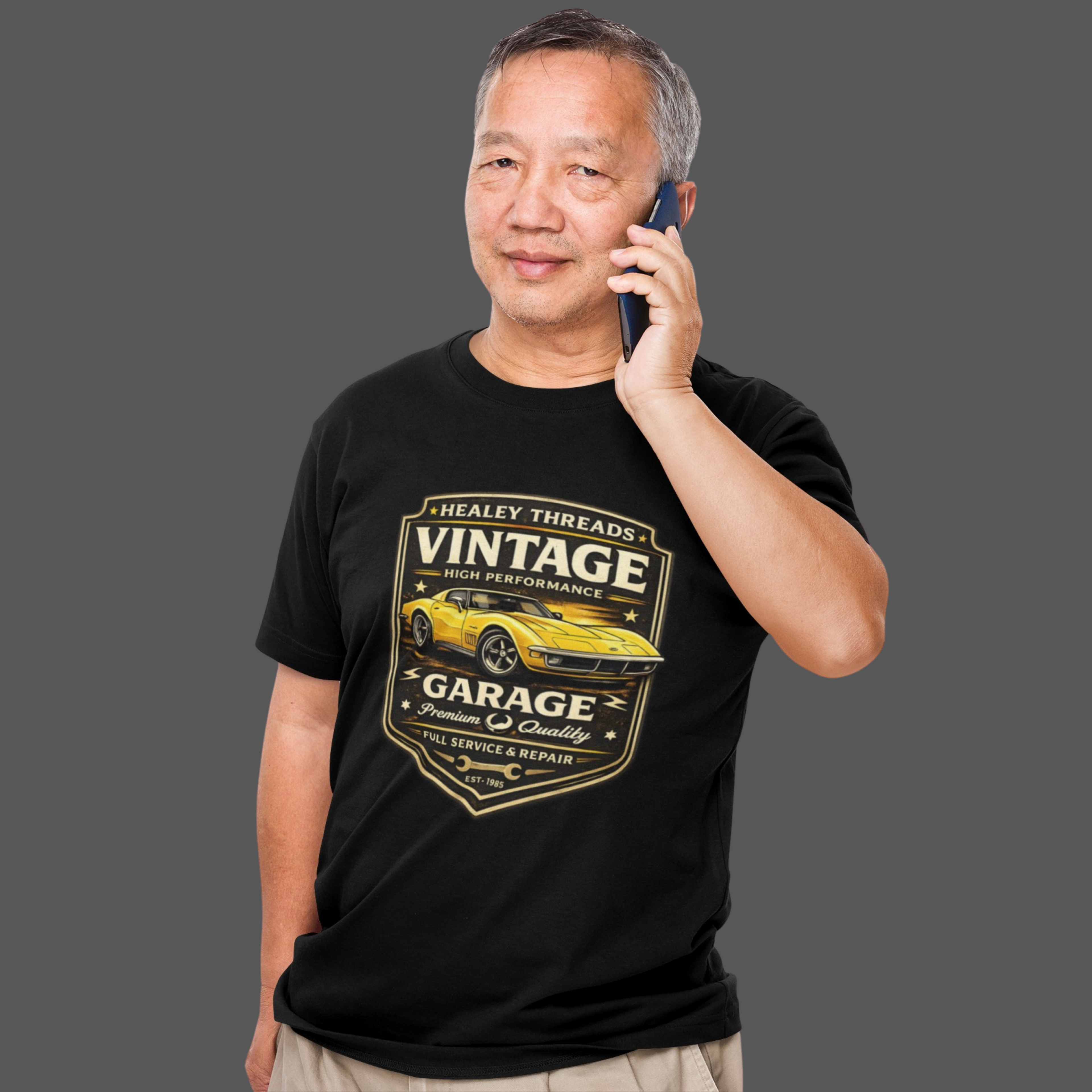 Man wearing a black t-shirt with a vintage classic car graphic, talking on a phone against a gray background