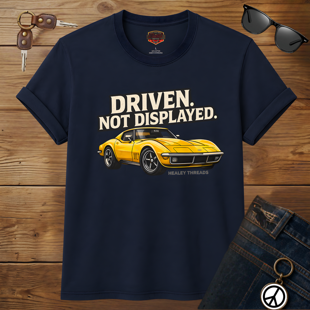 Navy blue t-shirt with yellow muscle car graphic and text 'Driven. Not Displayed.' on a wooden surface.