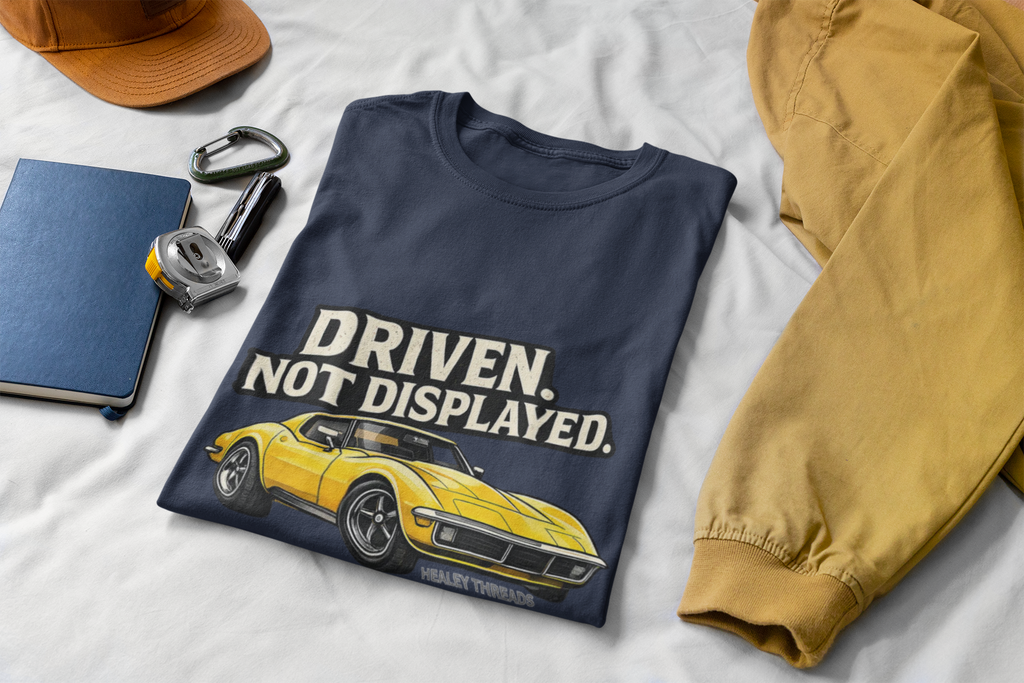Navy t-shirt with yellow muscle car graphic and 'Driven. Not Displayed.' text on a white surface with accessories.