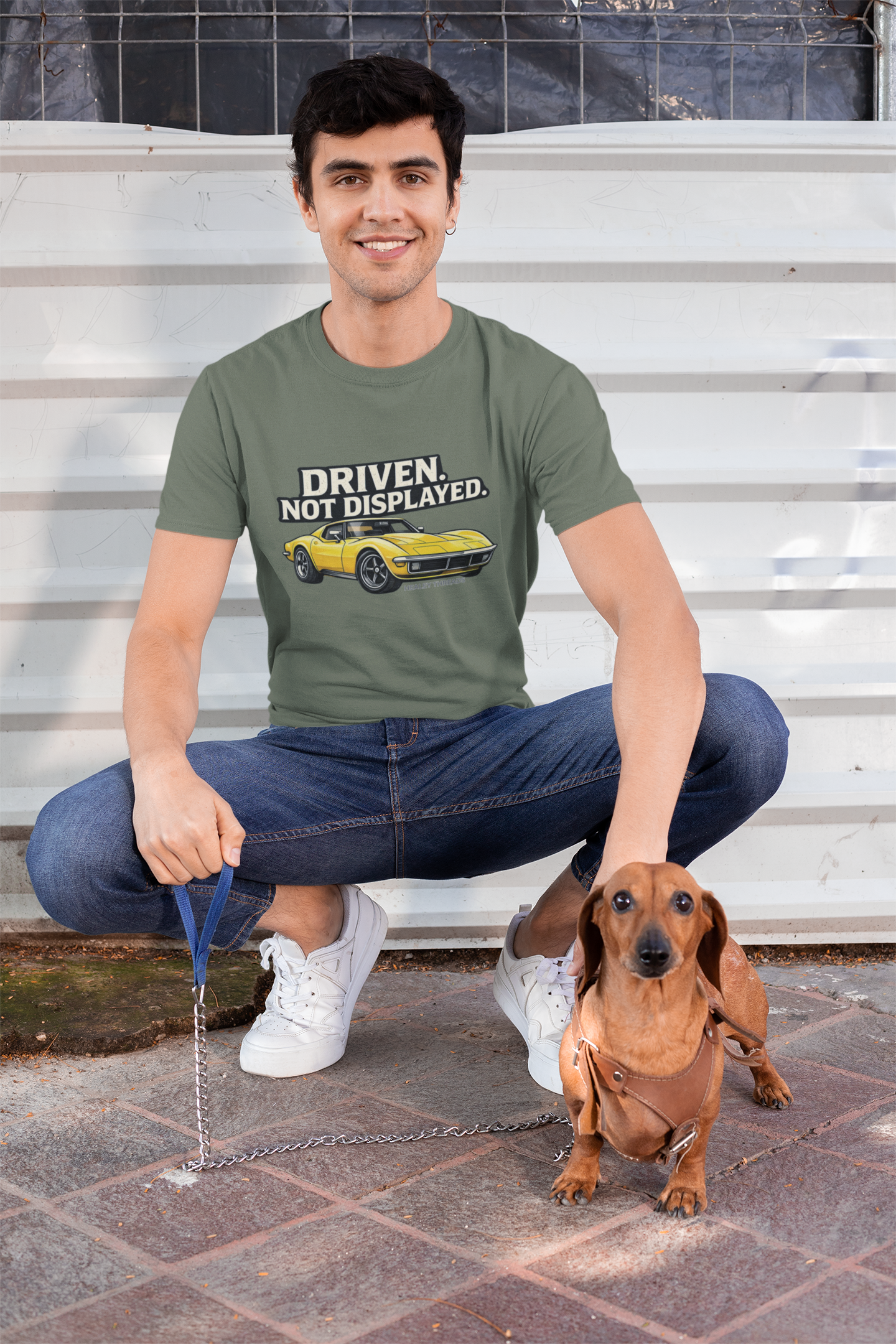 Man sitting on steps with a dachshund wearing a military green t-shirt with text and a muscle car graphic.
