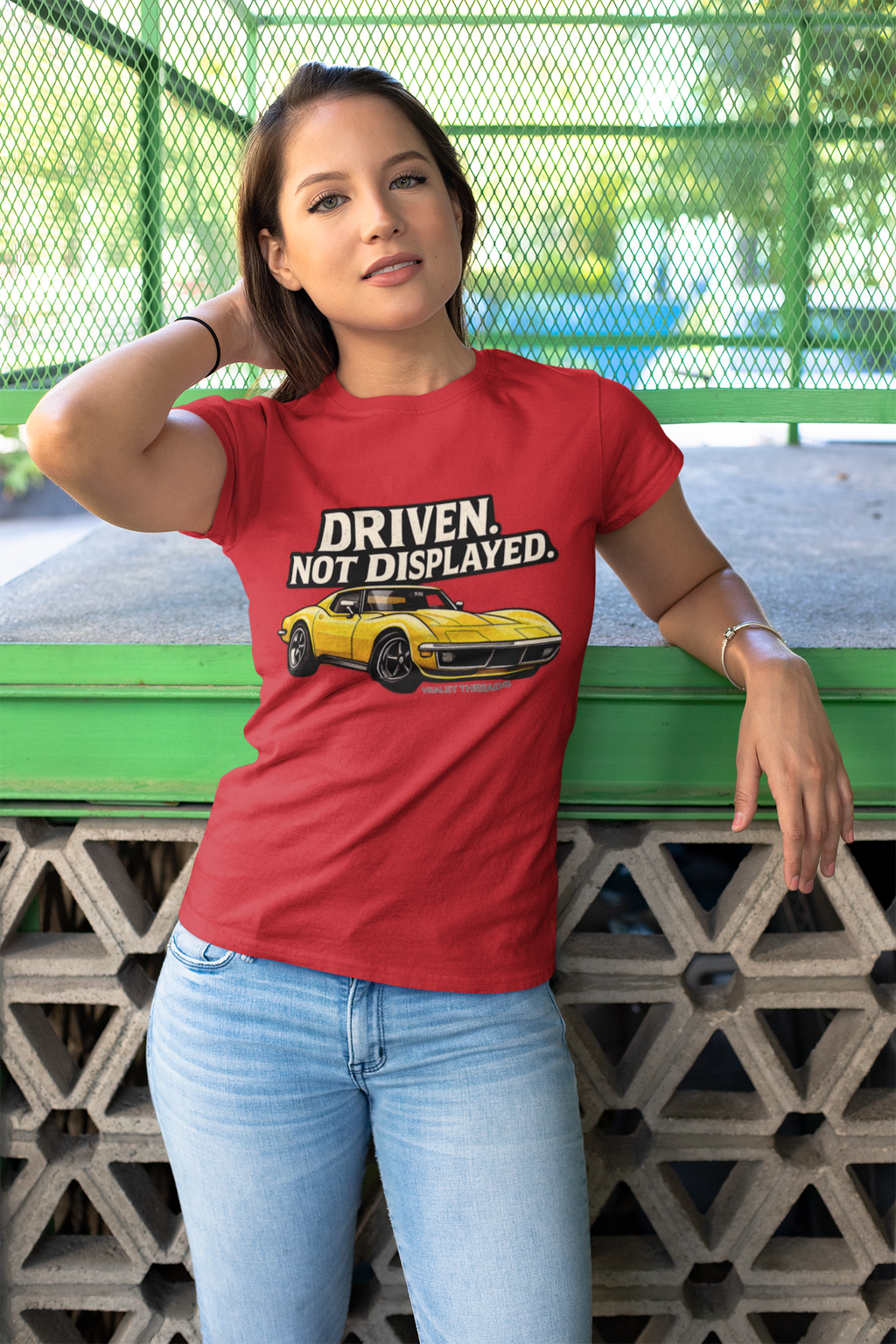 Woman wearing a red t-shirt with a graphic muscle car and text, standing against a green metal fence.