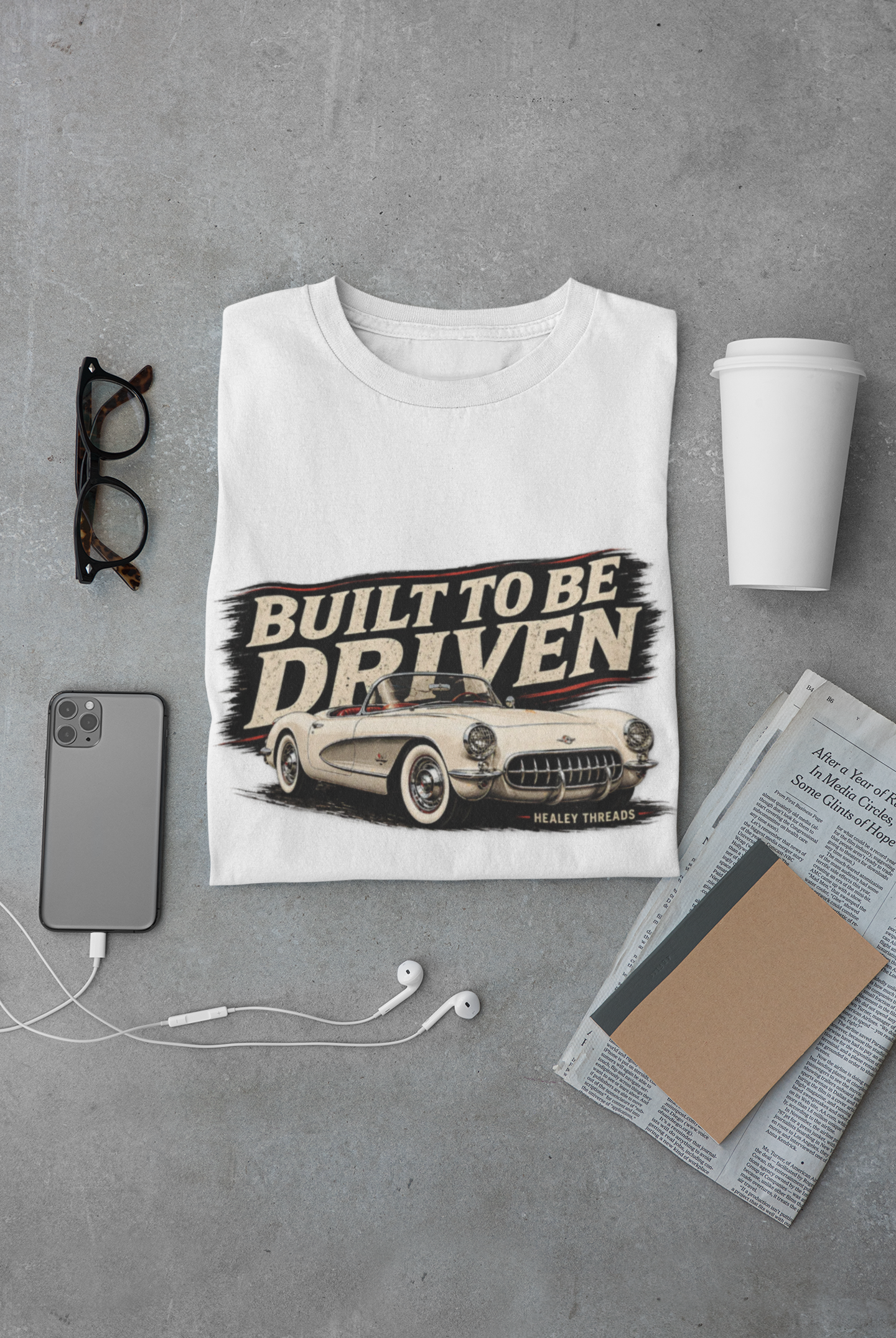 White t-shirt with 'Built to be Driven' graphic on a gray surface with phone, glasses, and newspaper.
