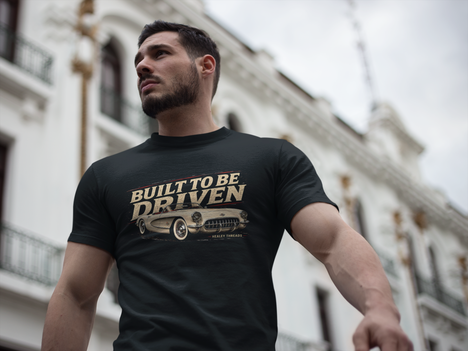 Man wearing a black t-shirt with a graphic built to be driven design and text, standing in front of a building.