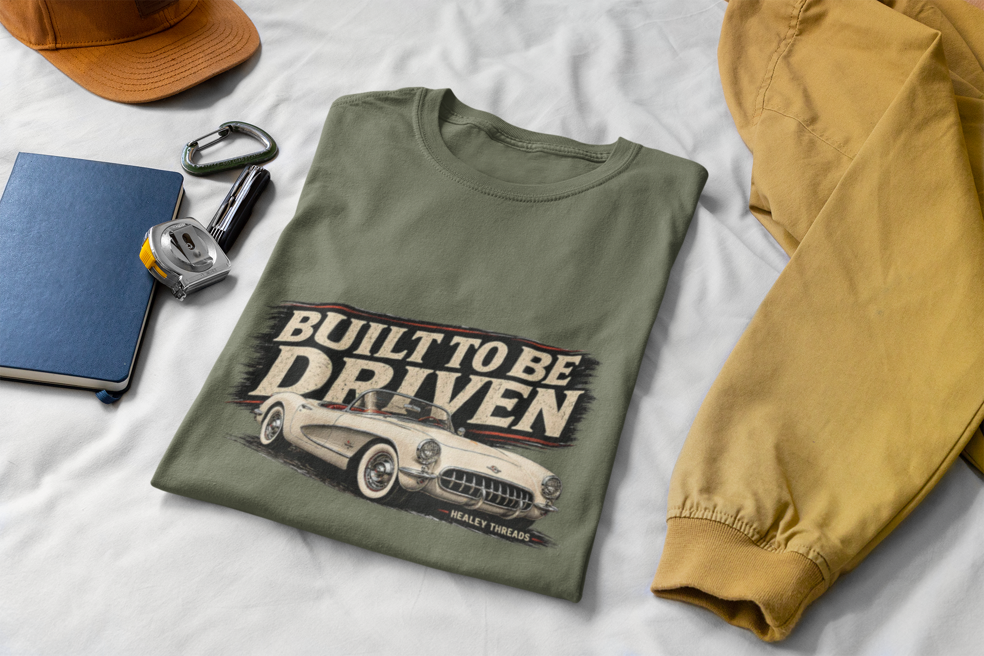 Military green t-shirt with 'Built to be Driven' graphic on a white surface with accessories.