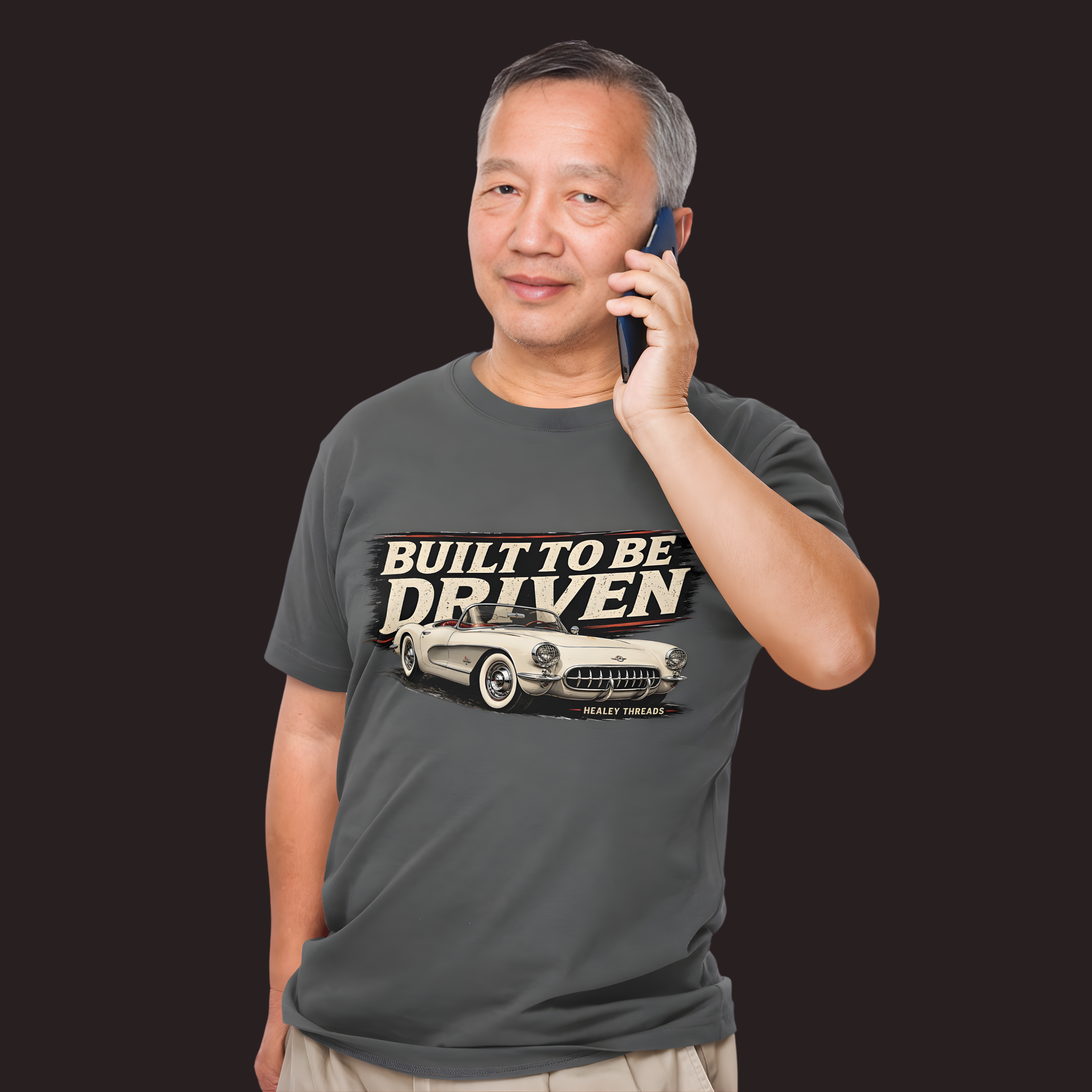 Man wearing a dark heather t-shirt with a classic car graphic and text, talking on a phone against a black background