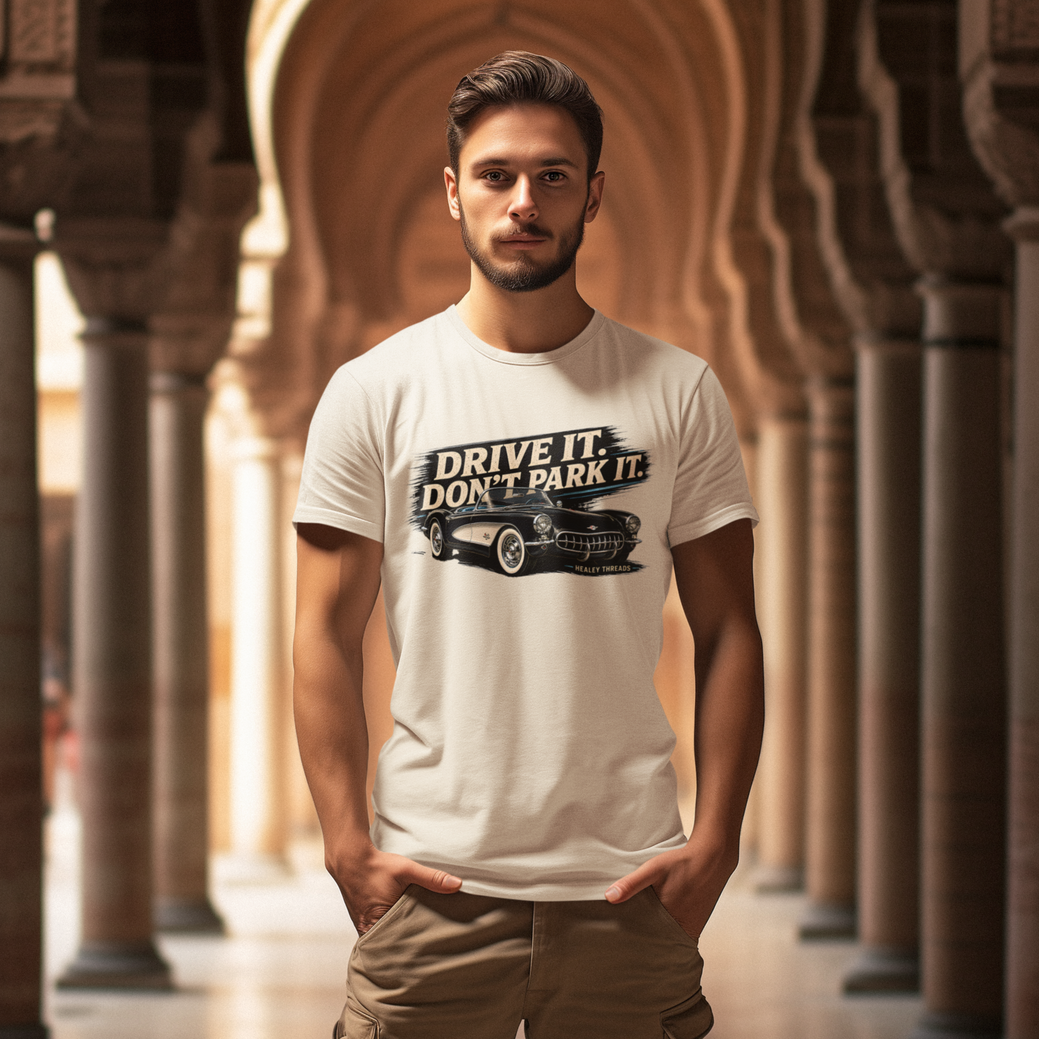 Man wearing a sand t-shirt with a vintage car graphic and text in an arched corridor.