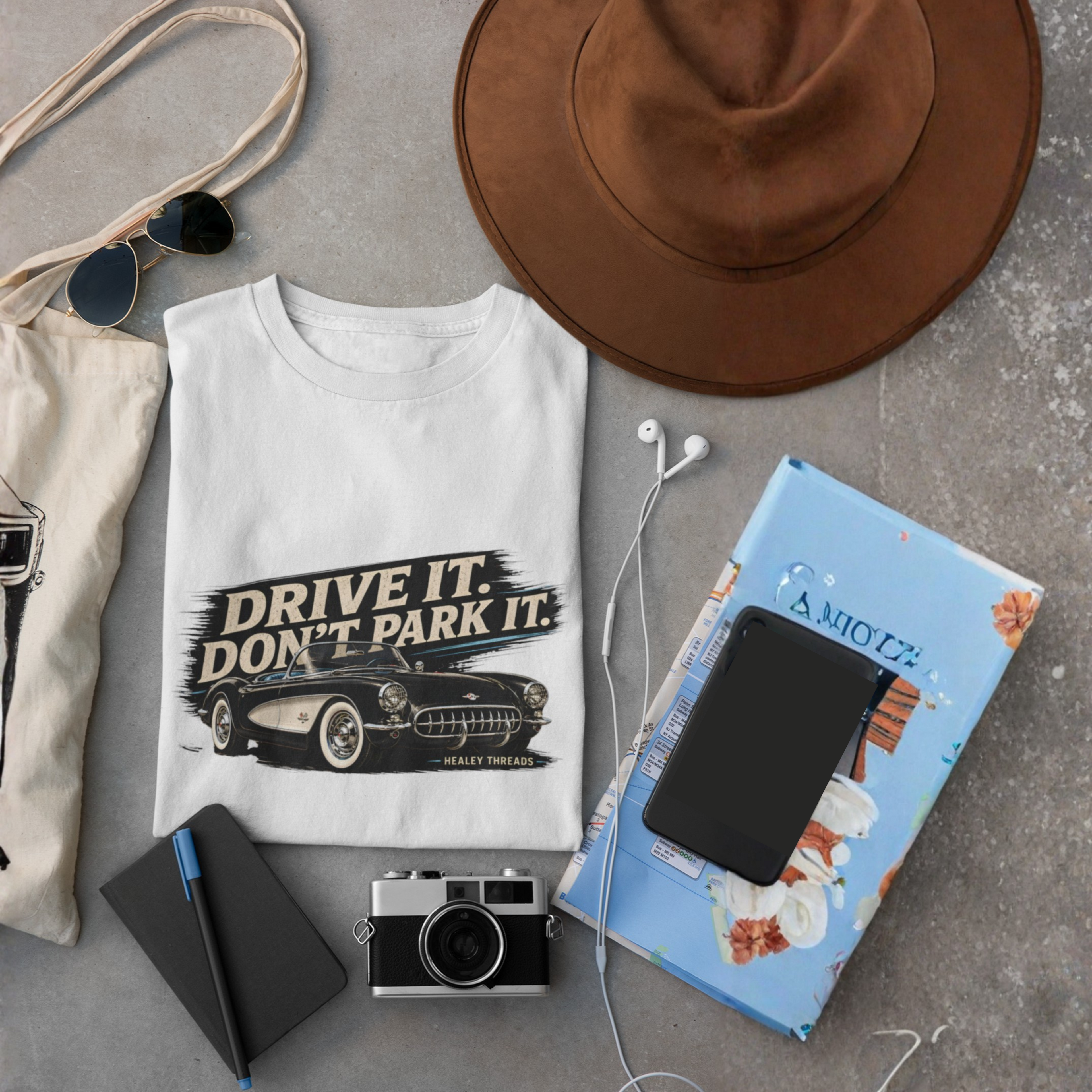 White t-shirt with graphic vintage car design, brown hat, camera, notebook, and sunglasses on a gray surface