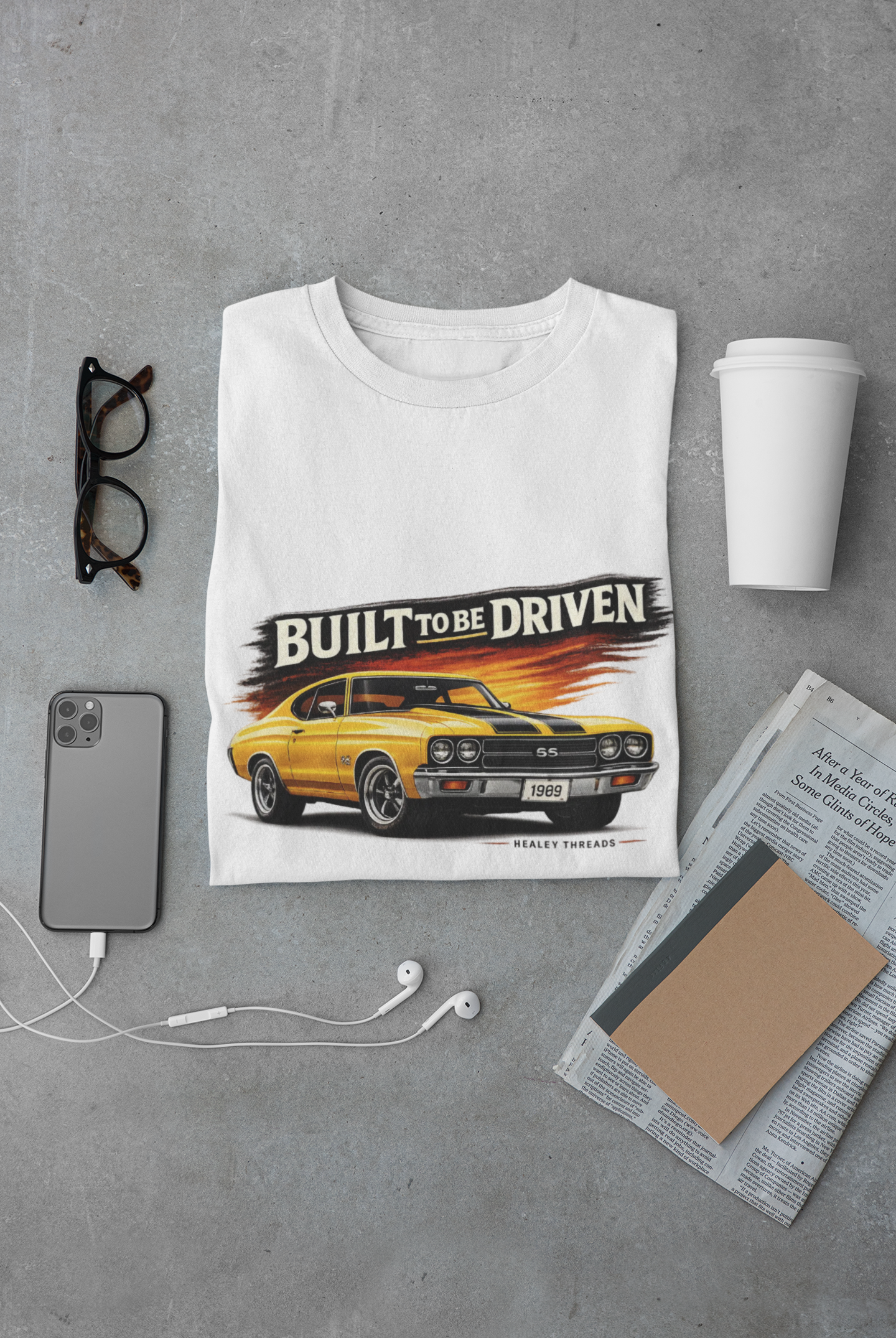 White t-shirt with a car graphic and 'Built to be Driven' text on a gray surface with a phone, glasses, and newspaper.