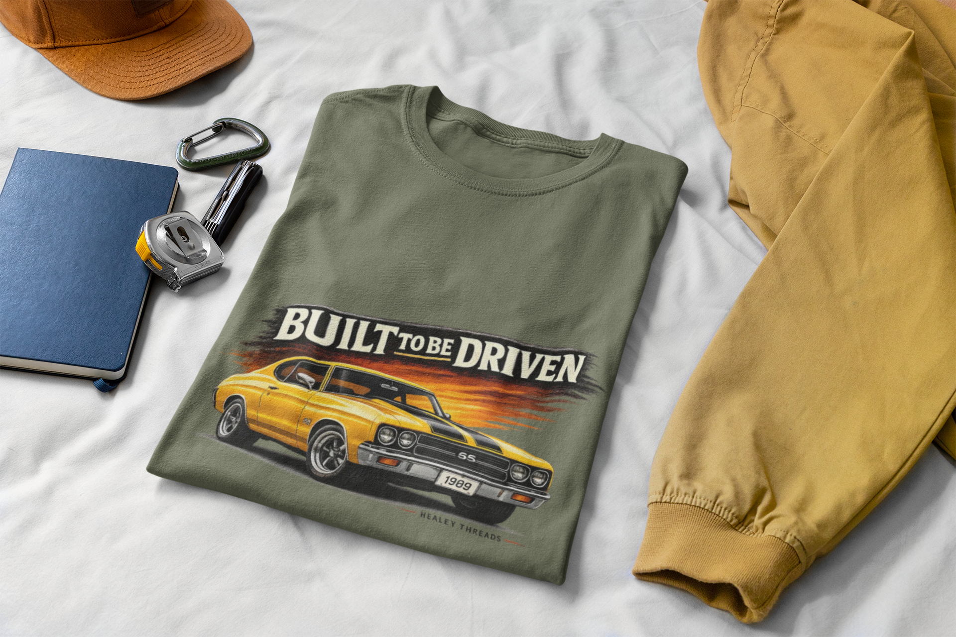 Military green t-shirt with car graphic and 'Built to be Driven' text on a white surface with accessories.