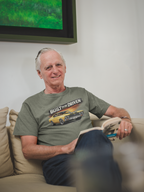 Man sitting on a couch wearing a military green t-shirt with a graphic car design, in a room with a television in the background.