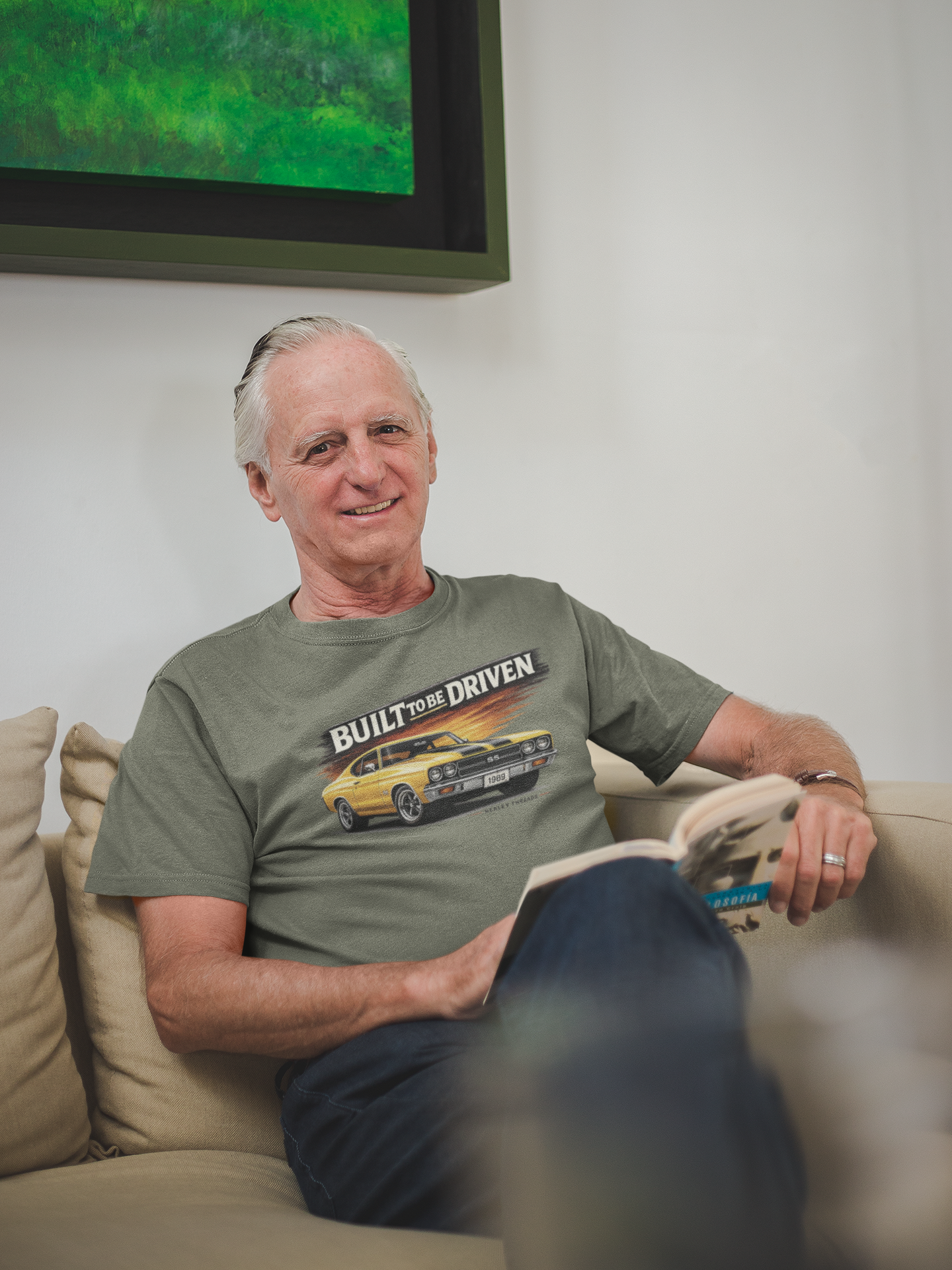 Man sitting on a couch wearing a military green t-shirt with a graphic car design, in a room with a television in the background.
