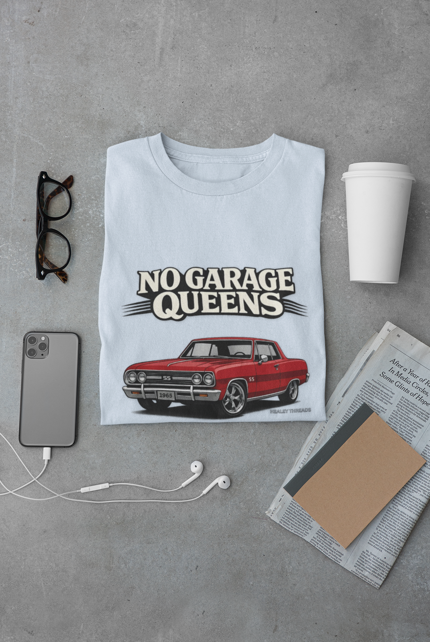 Light blue t-shirt with red car graphic and 'No Garage Queens' text on a gray surface with phone, glasses, and newspaper.