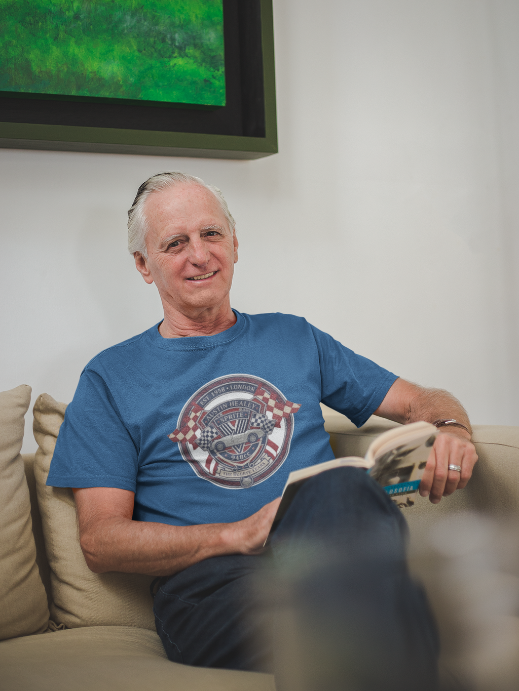 Man wearing a royal blue t-shirt with a graphic Healey Sprite design, sitting on a couch in a living room.