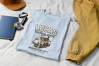 Light blue t-shirt with 'Lucas Electrical Works' design on a white surface with accessories.