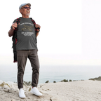Man standing on a sandy hill wearing a dark heather t-shirt with text and graphics, brown pants, white shoes, and a backpack.