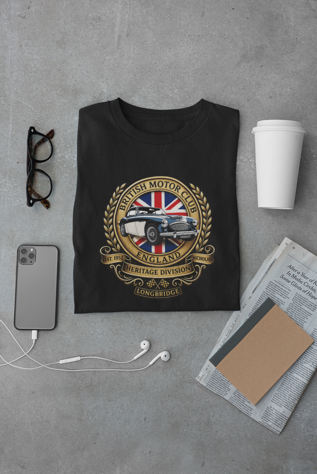 Black t-shirt with a British Motor Club logo on a gray surface with a phone, glasses, and coffee cup.